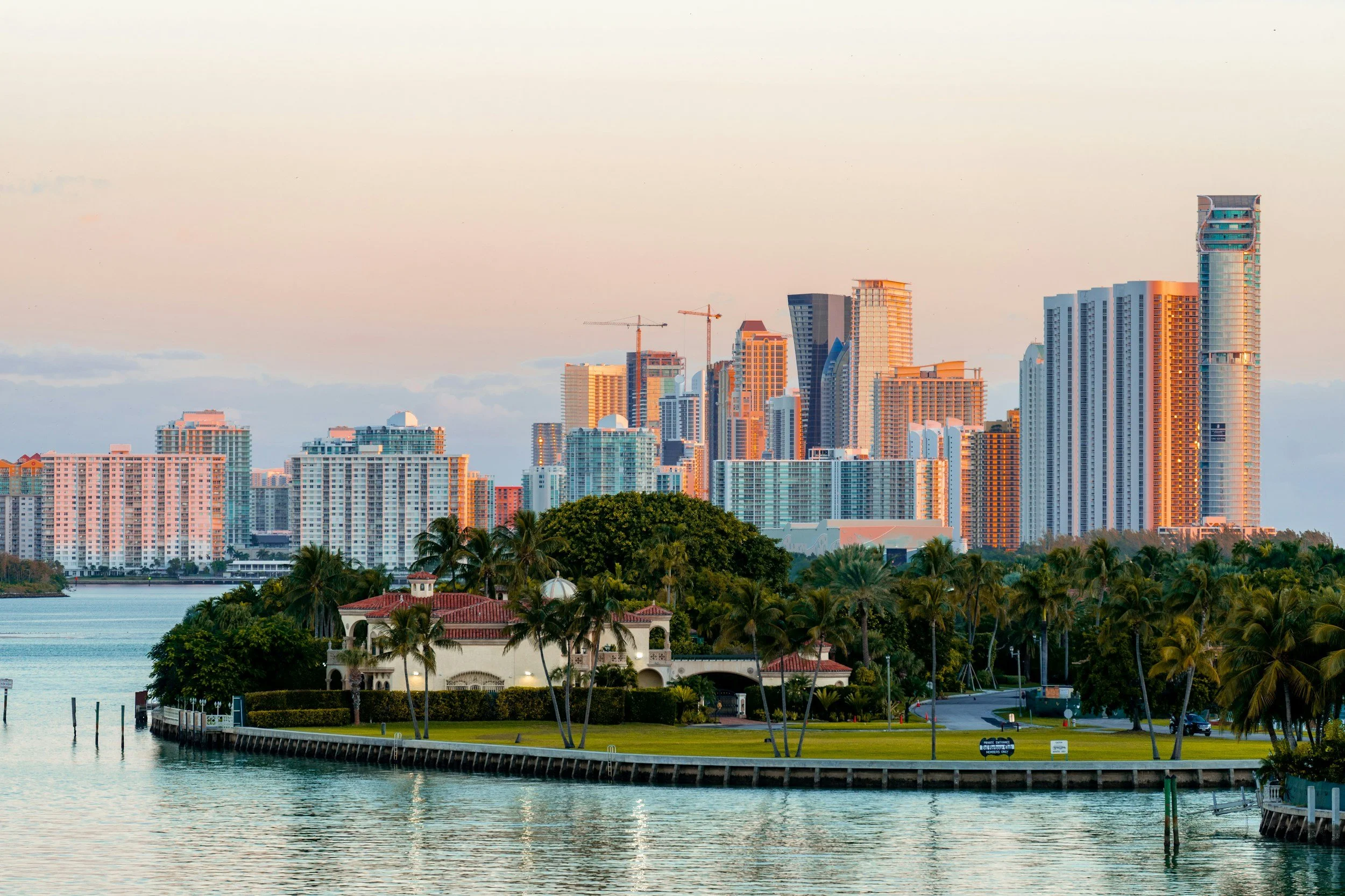 A city skyline with modern high-rise buildings viewed across a body of water, with lush greenery and a small historical-style building with a red roof in the foreground.