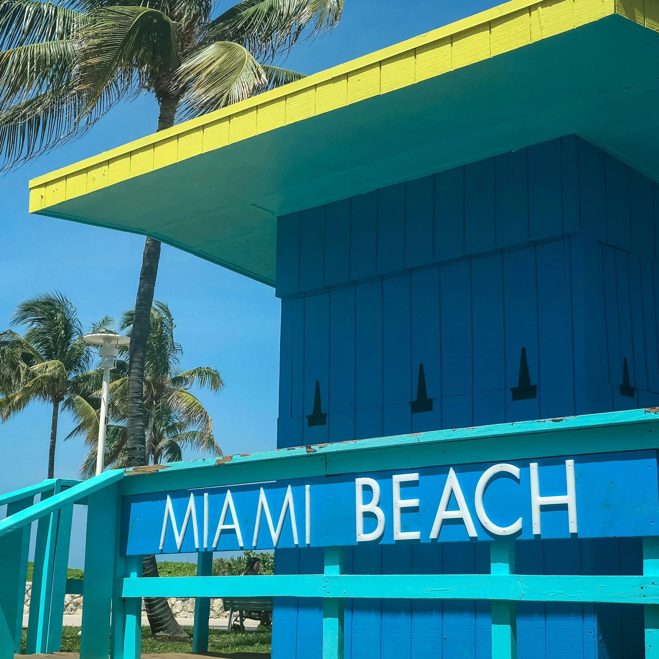 Colorful beach hut with a sign reading 'Miami Beach', surrounded by palm trees under a bright blue sky.