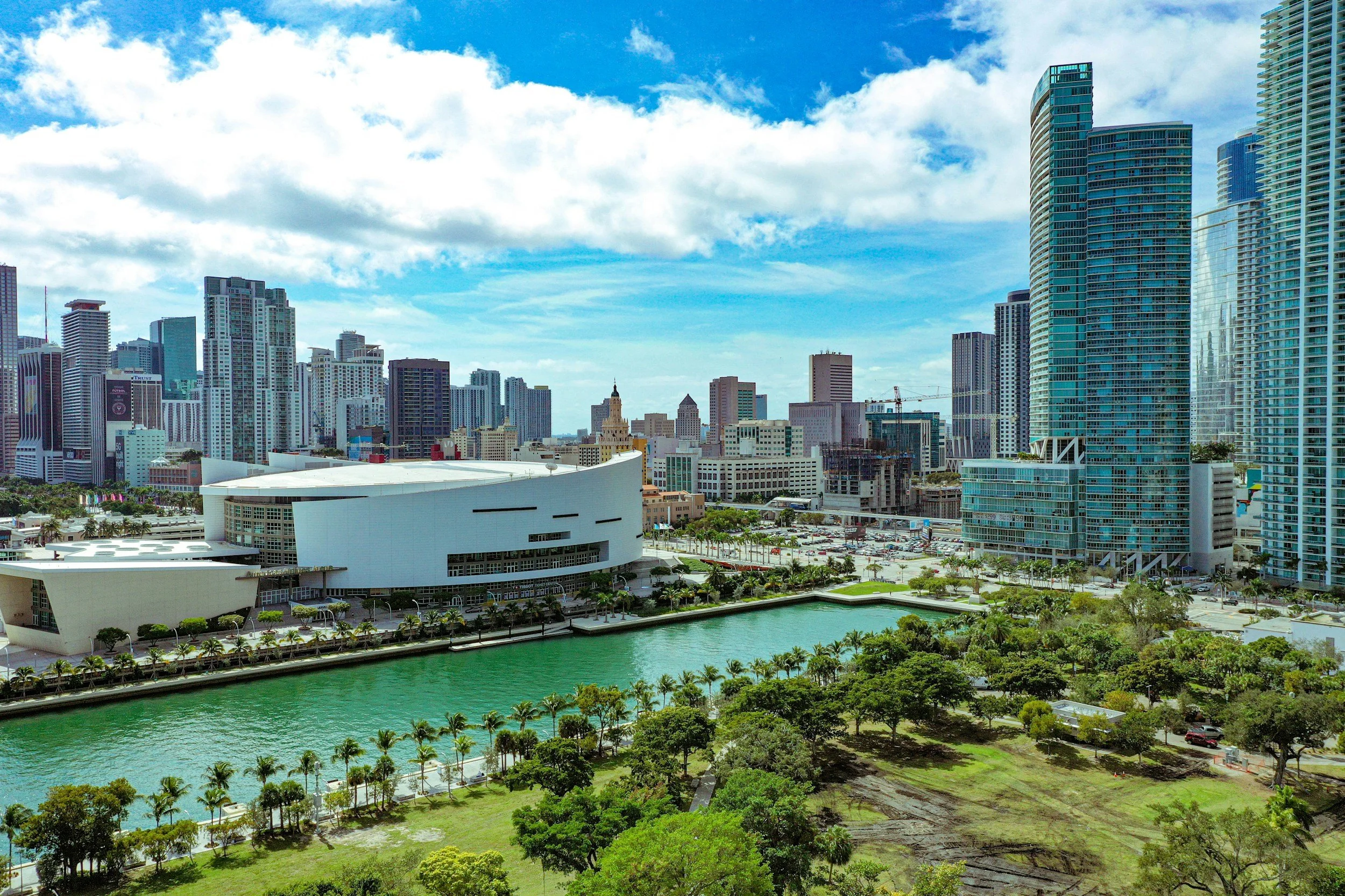 City skyline with modern skyscrapers, a body of water, and a park with trees and grass in the foreground.