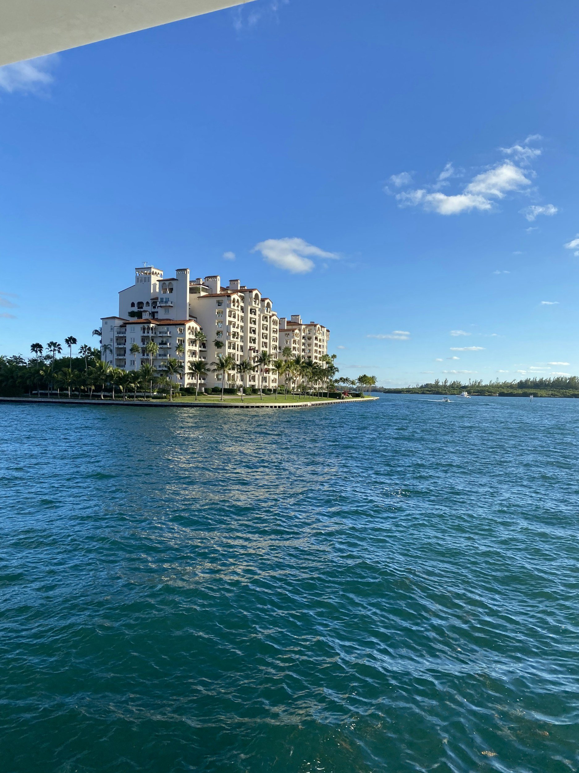 A large white building with multiple balconies situated on a water island, with a clear blue sky and a few clouds overhead.