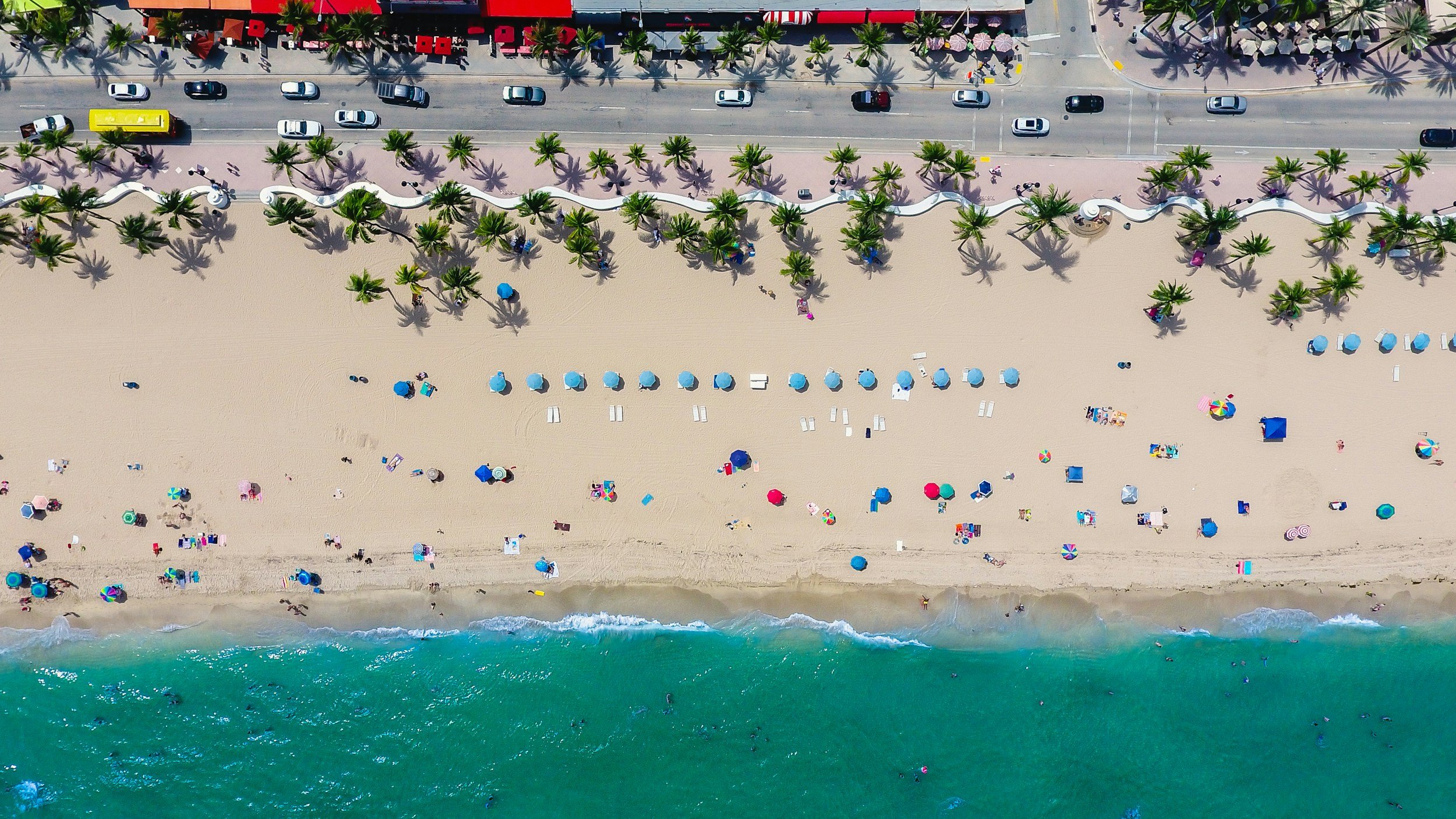 Aerial view of a beach with colorful umbrellas and lounge chairs, lined with palm trees along a promenade with cars and red umbrellas.