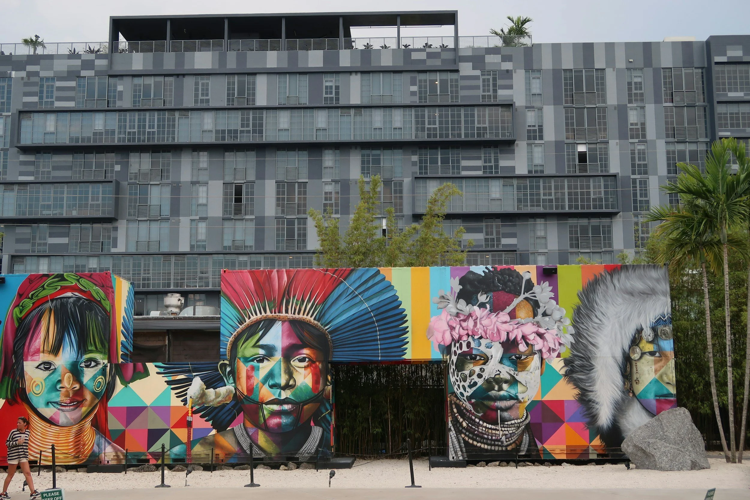 Colorful mural art depicting indigenous and cultural portraits on shipping containers in front of a modern high-rise building with trees and palm trees nearby.