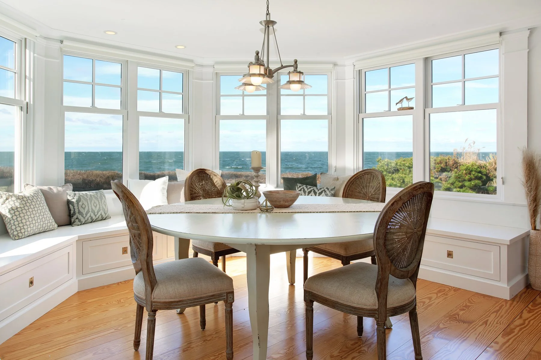 Dining room with a round table, four wooden chairs, built-in window seat with pillows, large windows showing ocean view, chandelier, and hardwood floor.