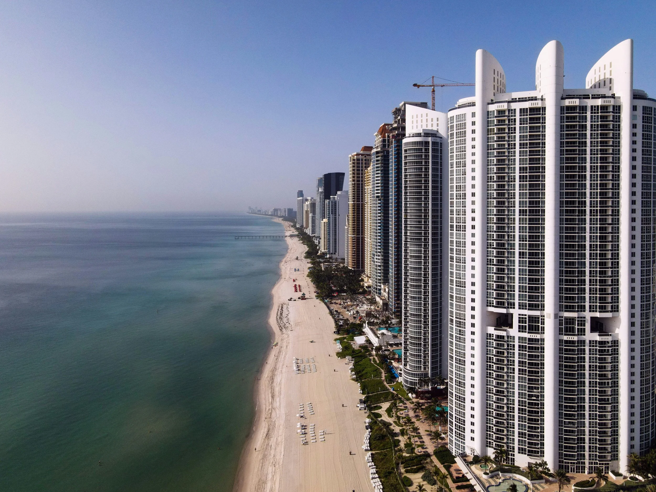Aerial view of high-rise buildings along a beach in Miami, Florida, showing sandy shoreline, ocean, clear sky, and construction crane on one of the buildings.