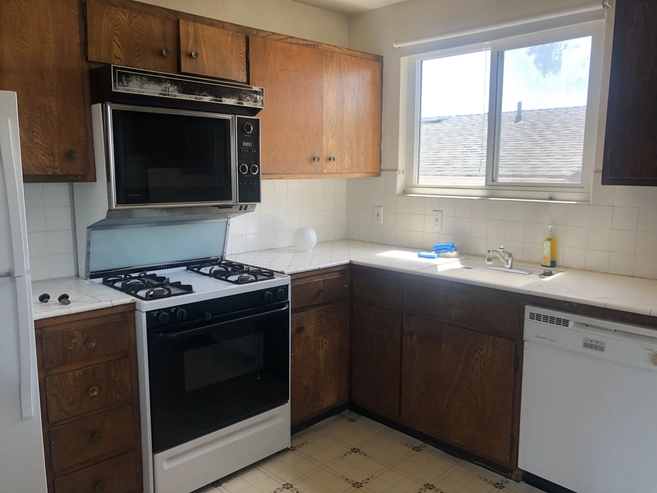 Kitchen with wooden cabinets, white gas stove, microwave oven, white tile countertops, double window above sink, white dishwasher, soap bottle, and blue dishcloths.