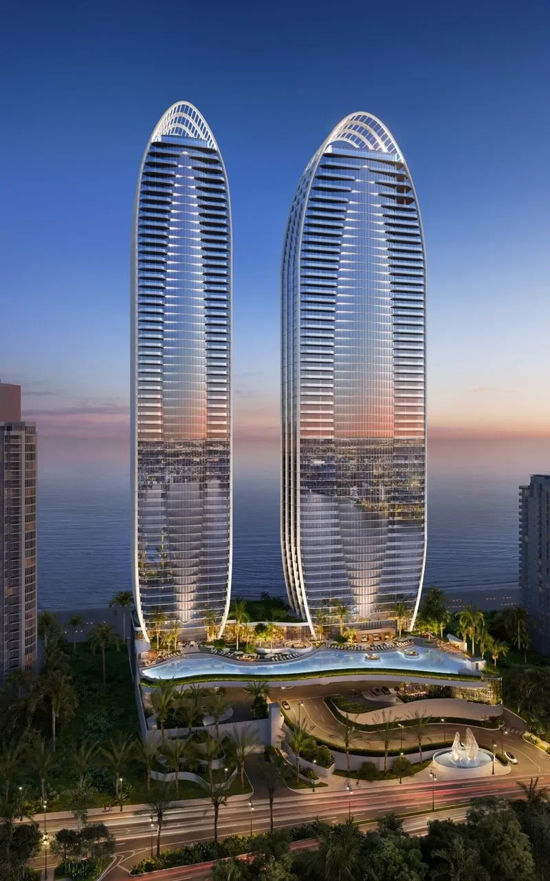 Twin modern high-rise buildings with glass facades reflecting sunset, overlooking the ocean, surrounded by green palms and a landscaped area with a pool and fountain.