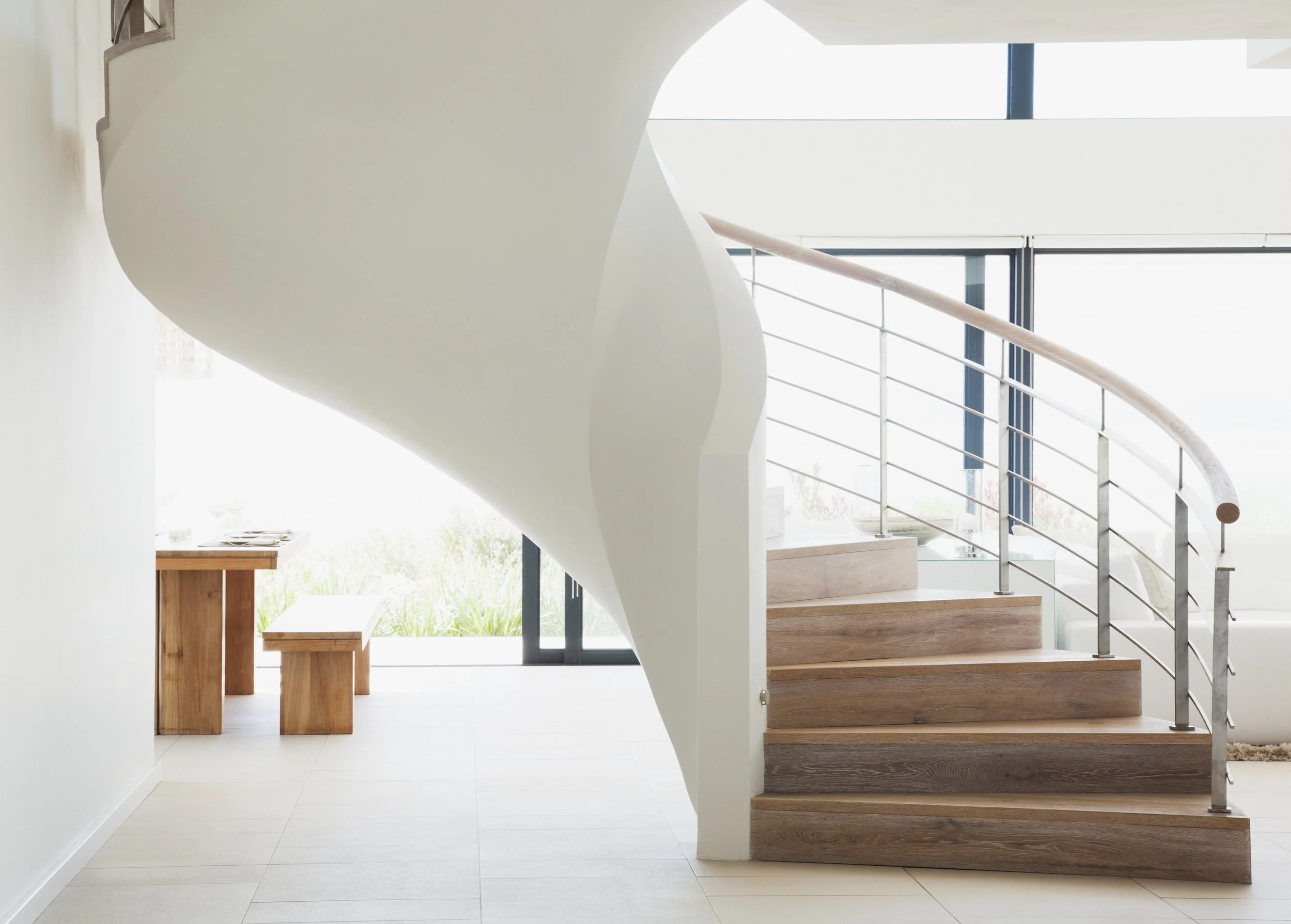 Modern interior with wooden staircase featuring metal handrail, white curved wall, and large floor-to-ceiling windows letting in natural light.