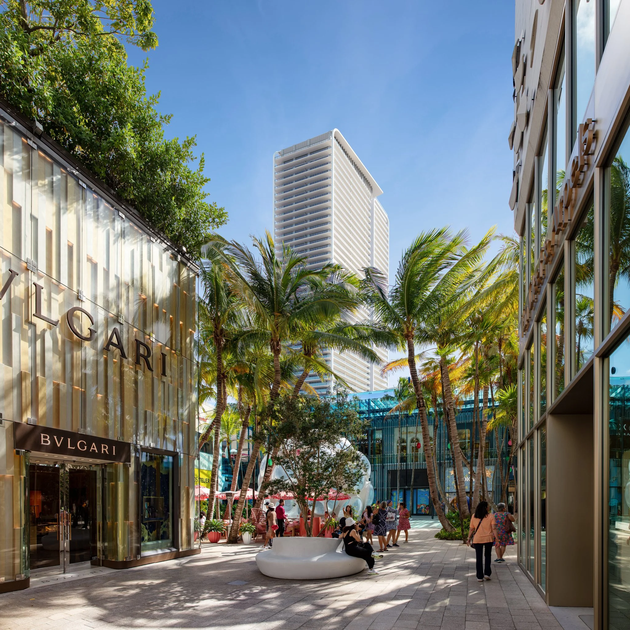 Shopping area with Bulgari store on the left, palm trees, people walking and sitting, and a tall modern building in the background under a clear blue sky.