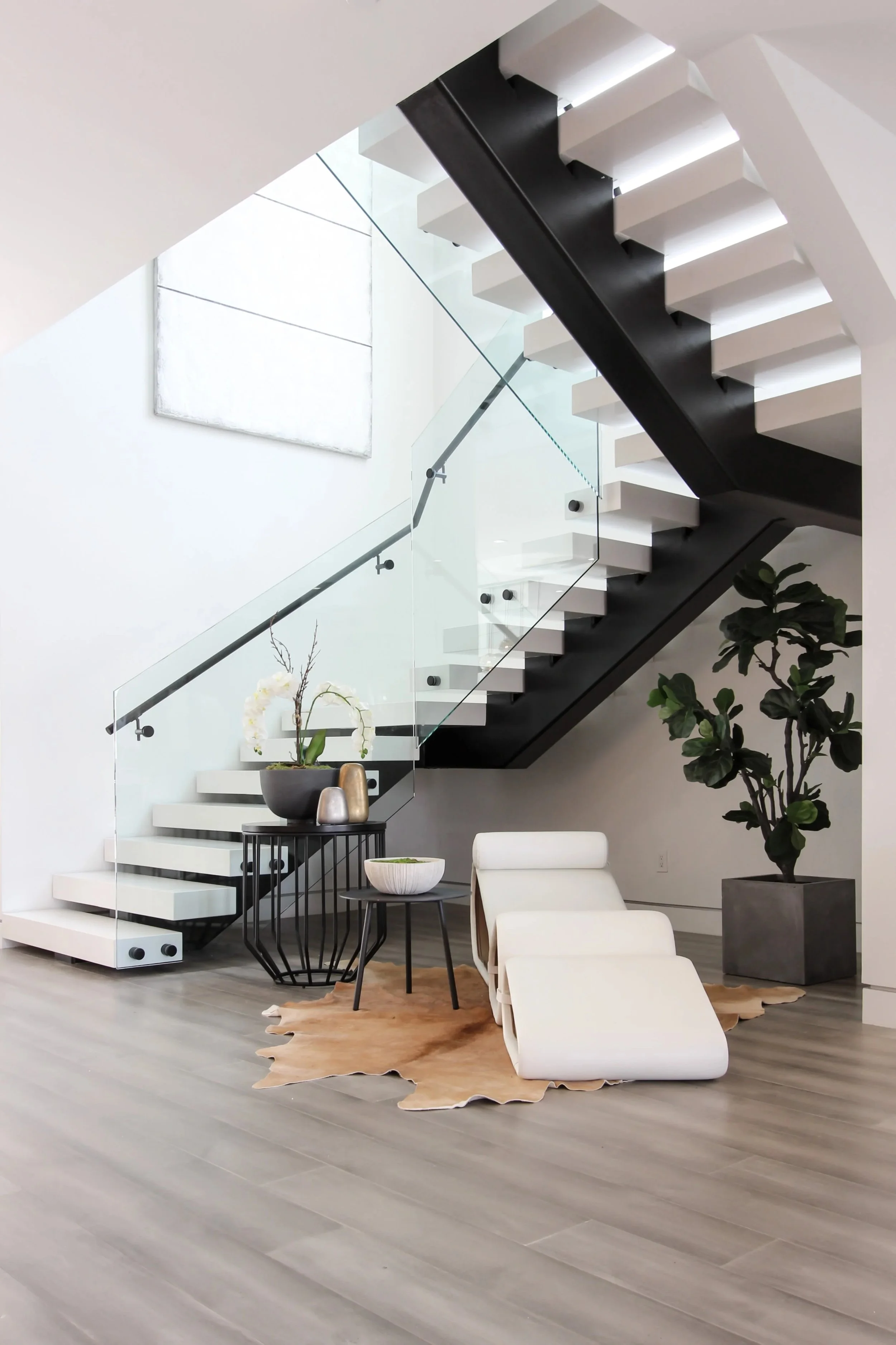 Modern staircase with glass railing, black and white steps, and a sitting area with a white chair, small black tables, a cowhide rug, a large plant, and decorative vases.