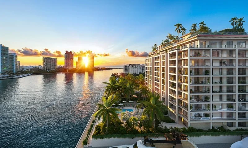 A waterfront view at sunset showing a high-rise building with balconies and palm trees on the roof, a pool, and a nearby city skyline across a river.