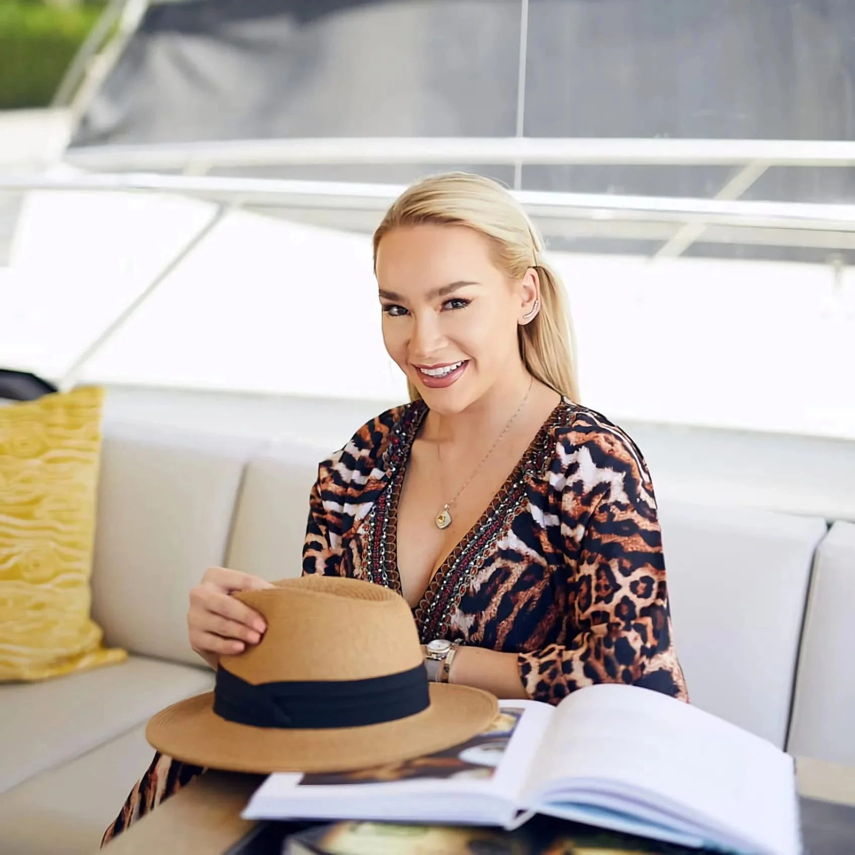 A smiling blonde woman wearing a leopard print top sitting on a white couch, holding a straw hat, with an open magazine on the table in front of her.