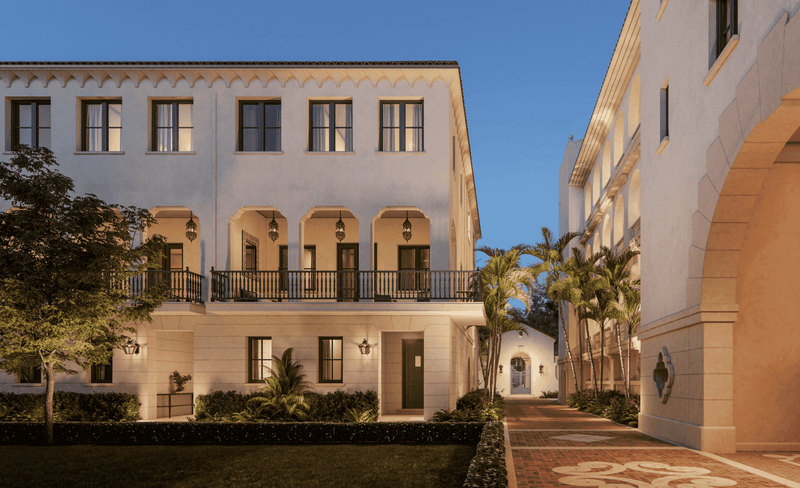 A white Mediterranean-style building with arched windows and balconies, palm trees, and a brick-paved walkway in the evening.