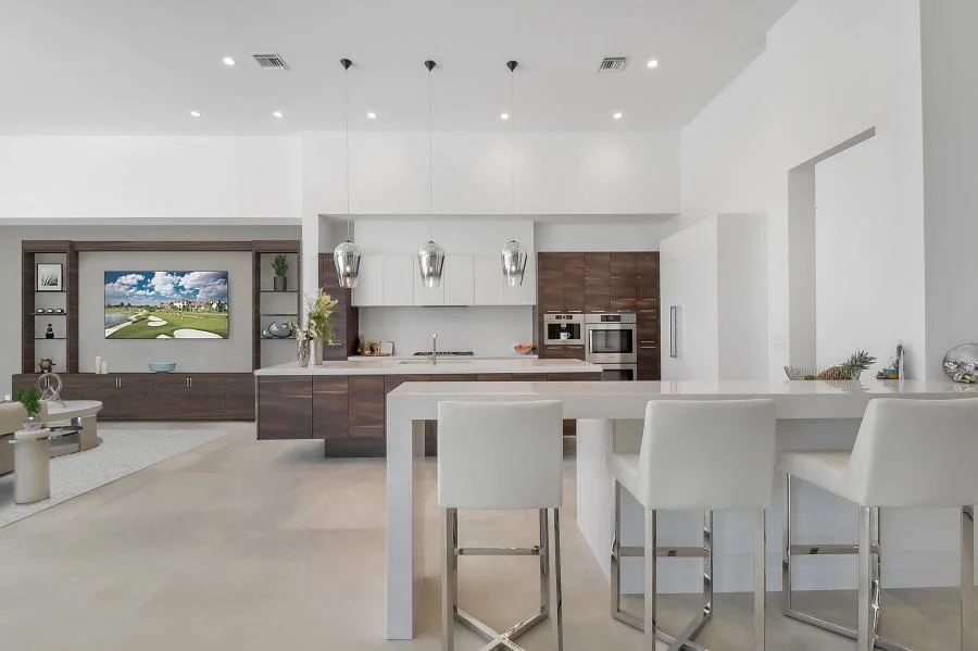 Modern open-concept kitchen and living room with white walls, brown cabinets, a TV, and a large white breakfast bar with white chairs.