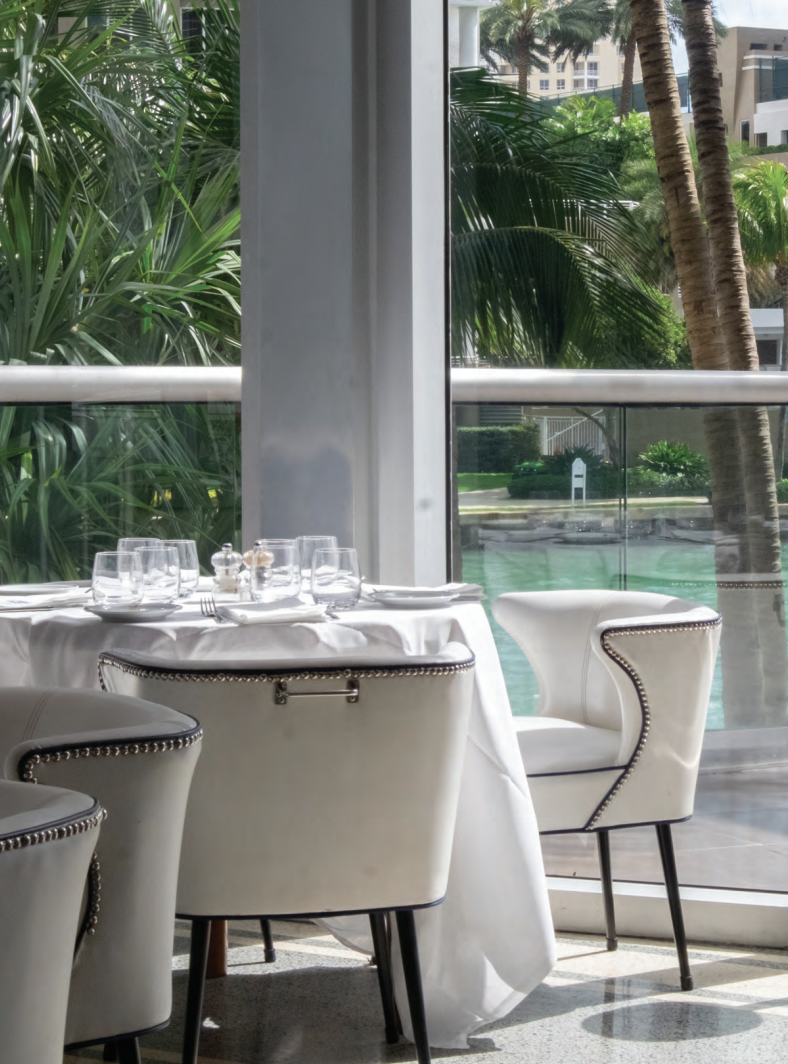 Indoor dining table set with white tablecloth, glassware, and silverware near large window with outdoor view of palm trees and a swimming pool.