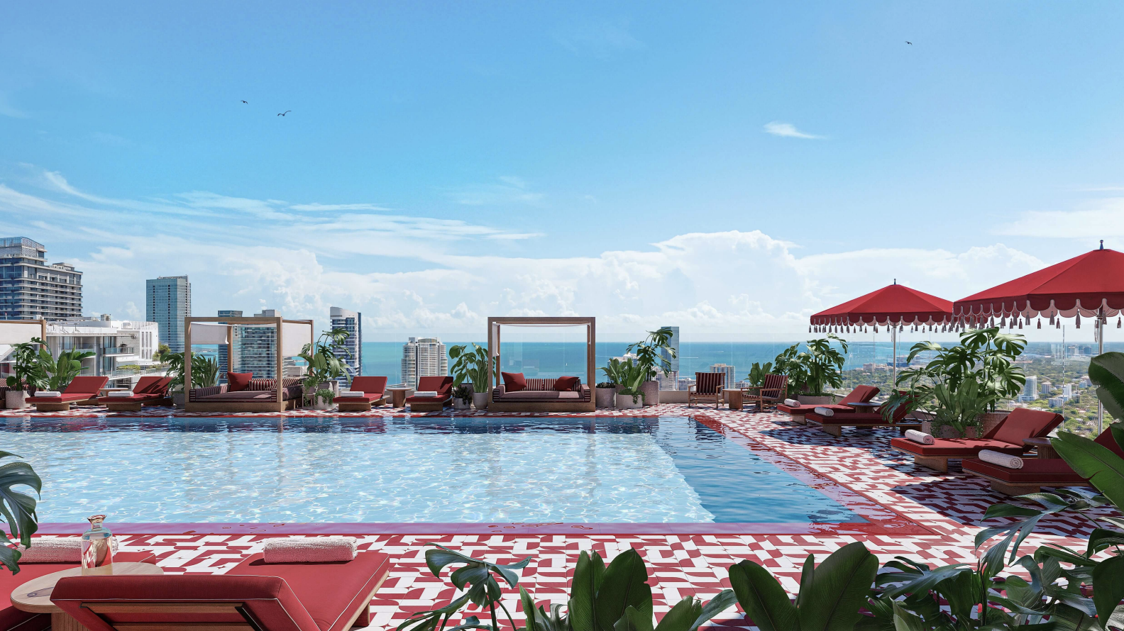 Rooftop pool area with lounge chairs, umbrellas, and potted plants overlooking a city skyline and ocean under a blue sky.