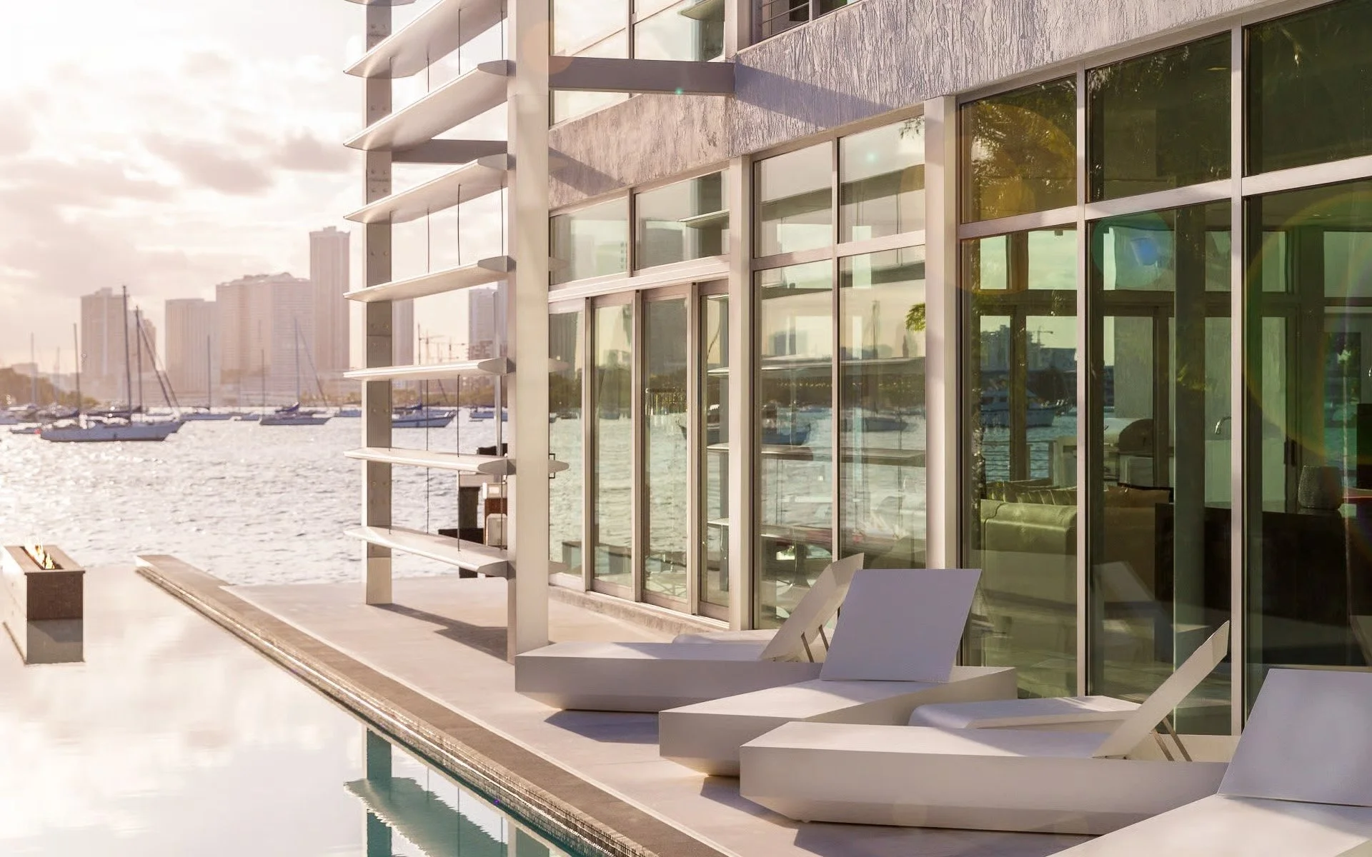 Modern hotel balcony with lounge chairs, overlooking a river with sailboats and a city skyline at sunset.