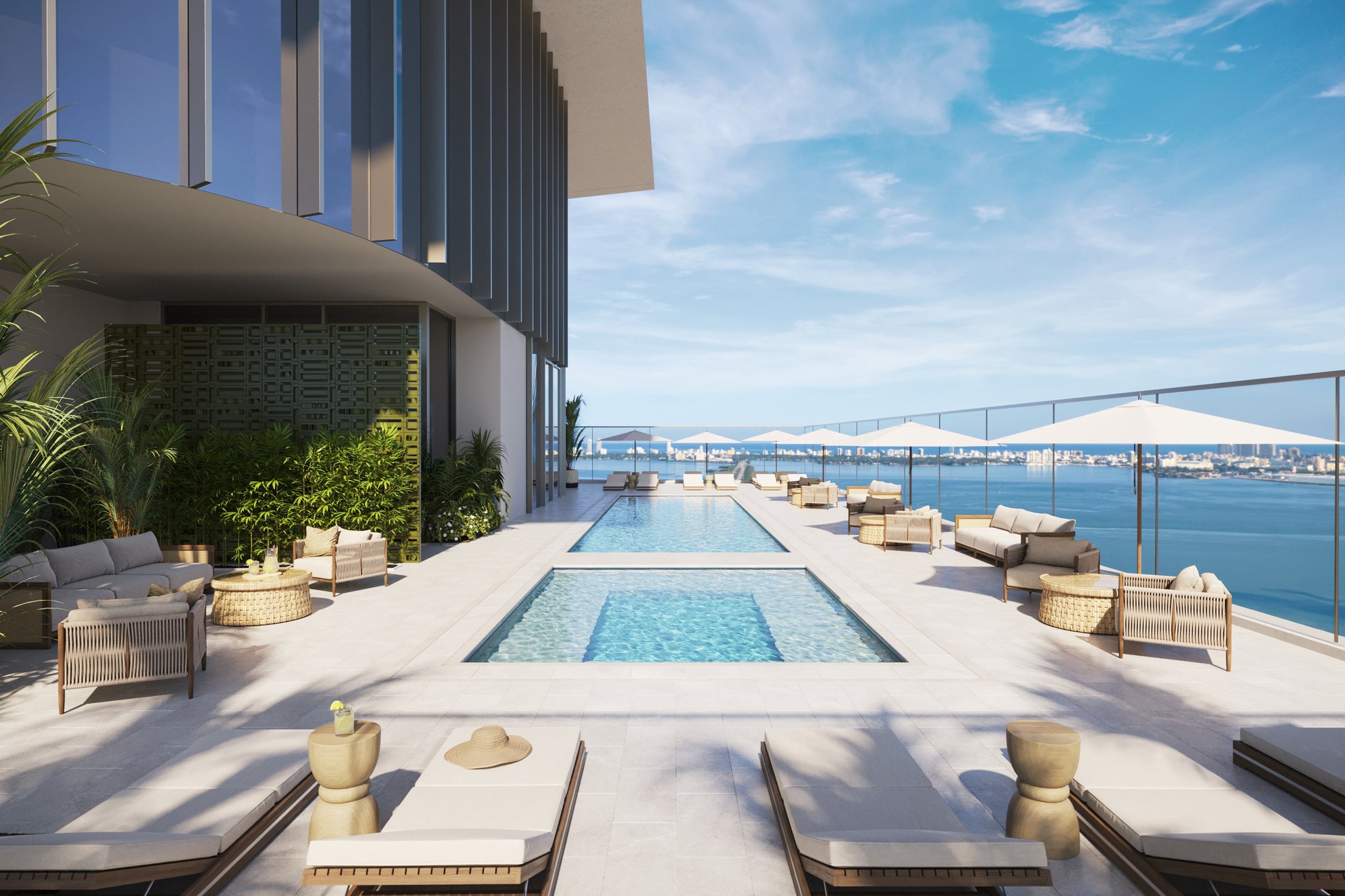 Luxury poolside terrace with lounge chairs, umbrellas, and views of a body of water and city skyline in the distance.