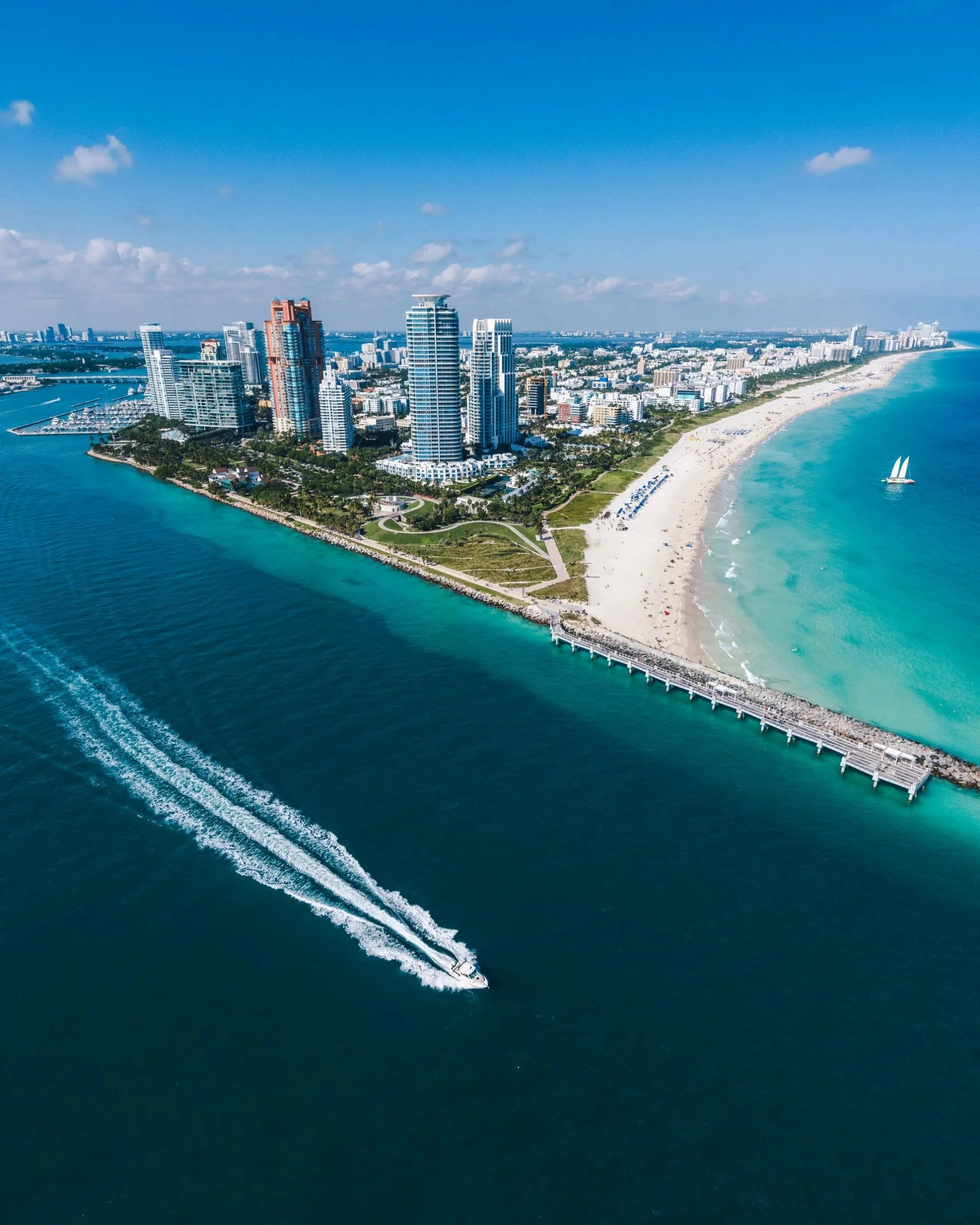Aerial view of a coastal city with tall modern buildings, a sandy beach, a pier, and a boat with sails on turquoise water.