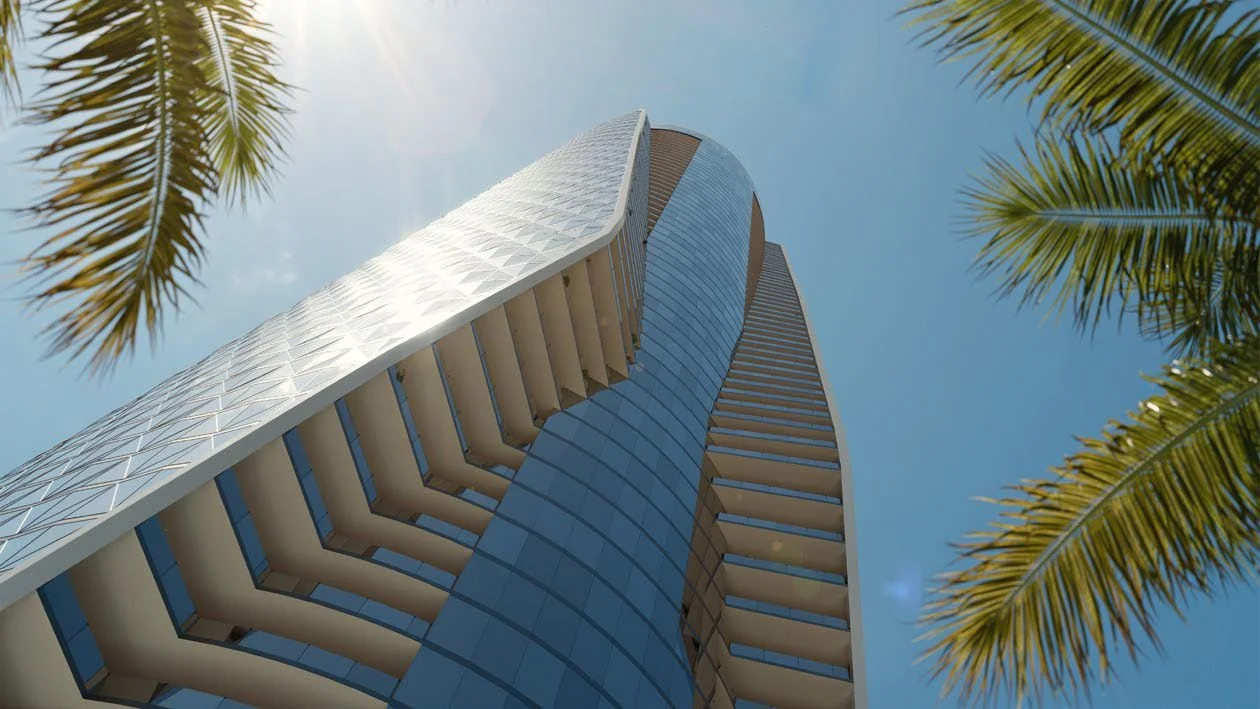 Looking up at a modern glass skyscraper with rounded edges, framed by palm trees against a clear blue sky.