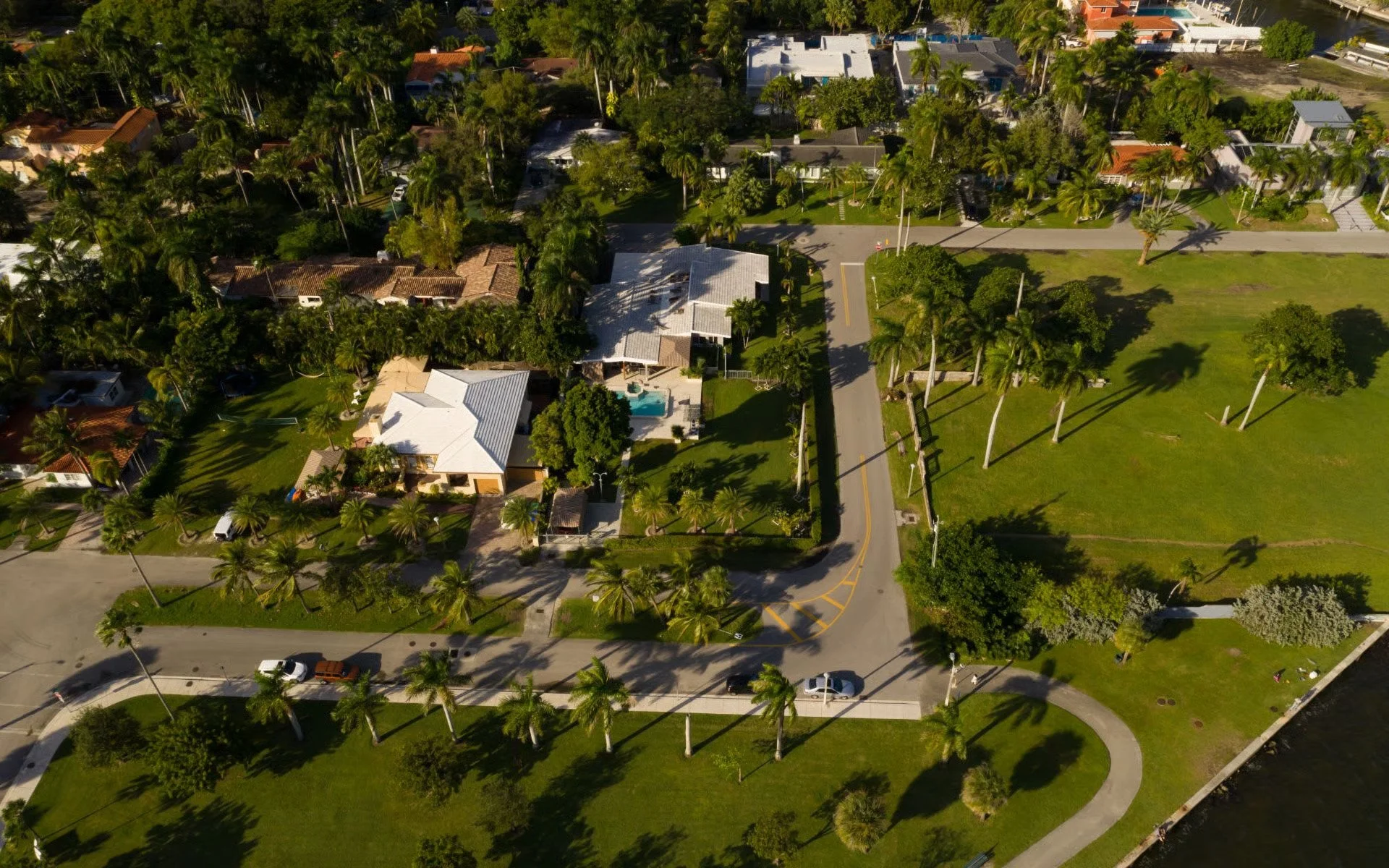 Aerial view of a residential neighborhood with houses, palm trees, and a large open grassy park area near a water body.