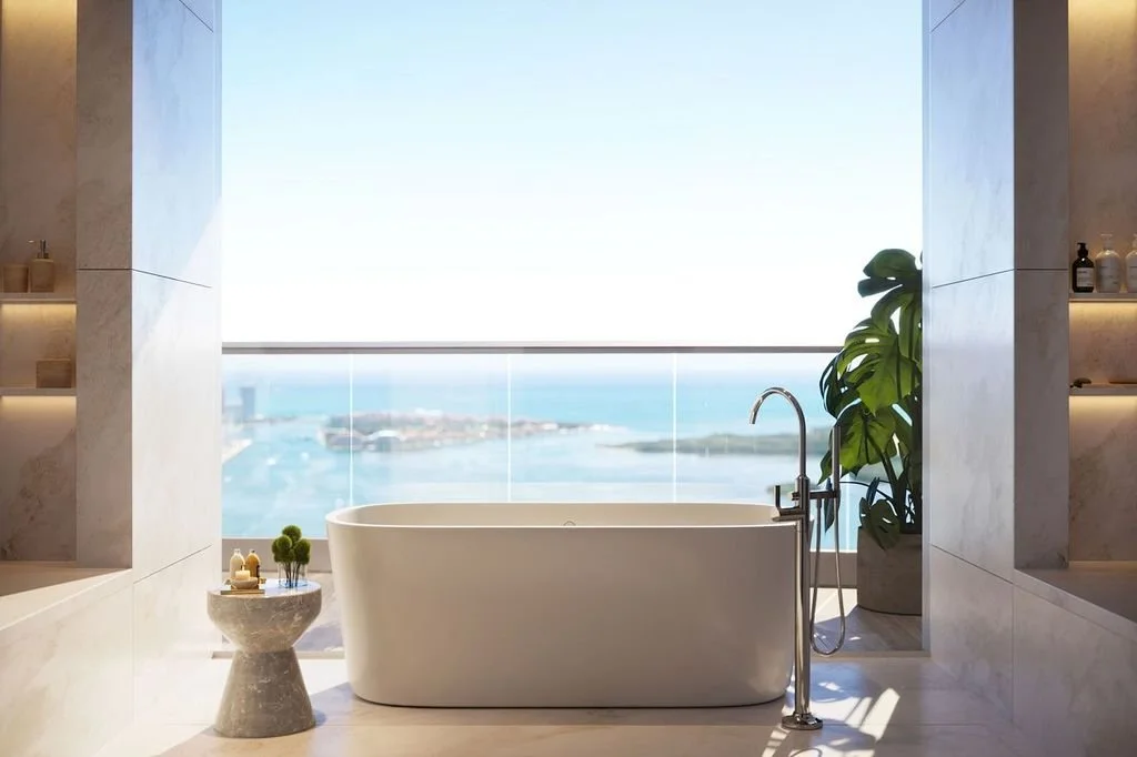 Modern bathroom with a freestanding bathtub facing a large window with ocean view, potted plant beside the tub, and a small stone side table with toiletries.