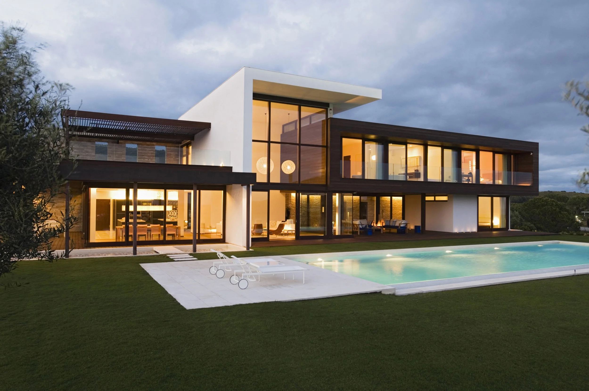 Modern two-story house with large glass windows, illuminated interior, swimming pool, and a well-manicured lawn during evening.