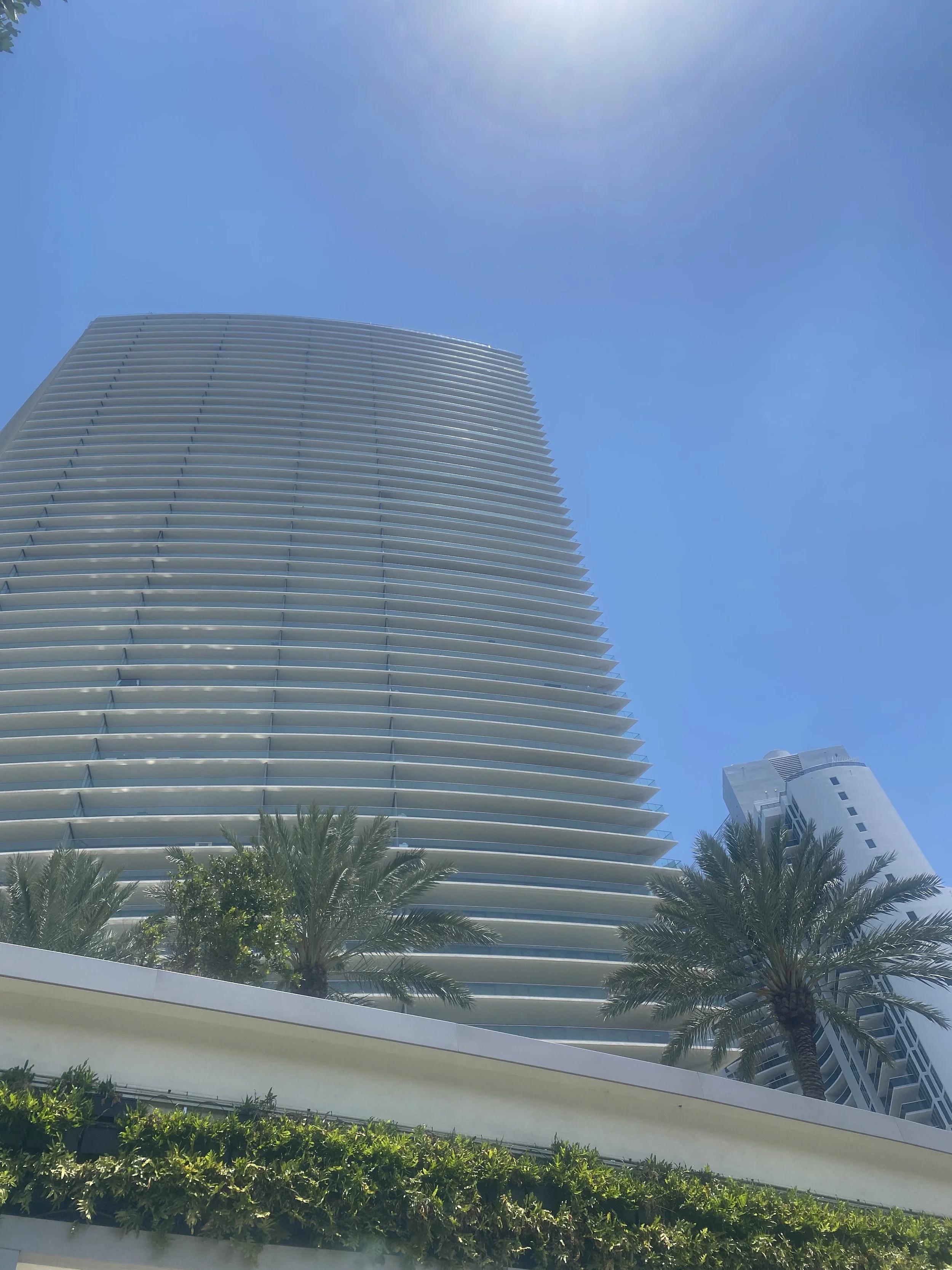 A tall modern skyscraper with horizontal white slats, blue sky background, and palm trees at the base.