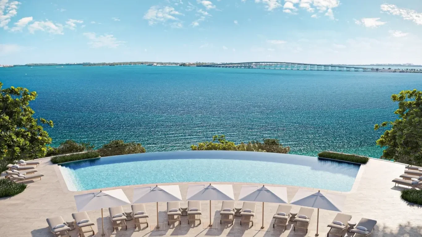 Luxury infinity pool overlooking a large body of water, with several white lounge chairs and umbrellas on a paved deck, surrounded by green trees, under a partly cloudy sky.
