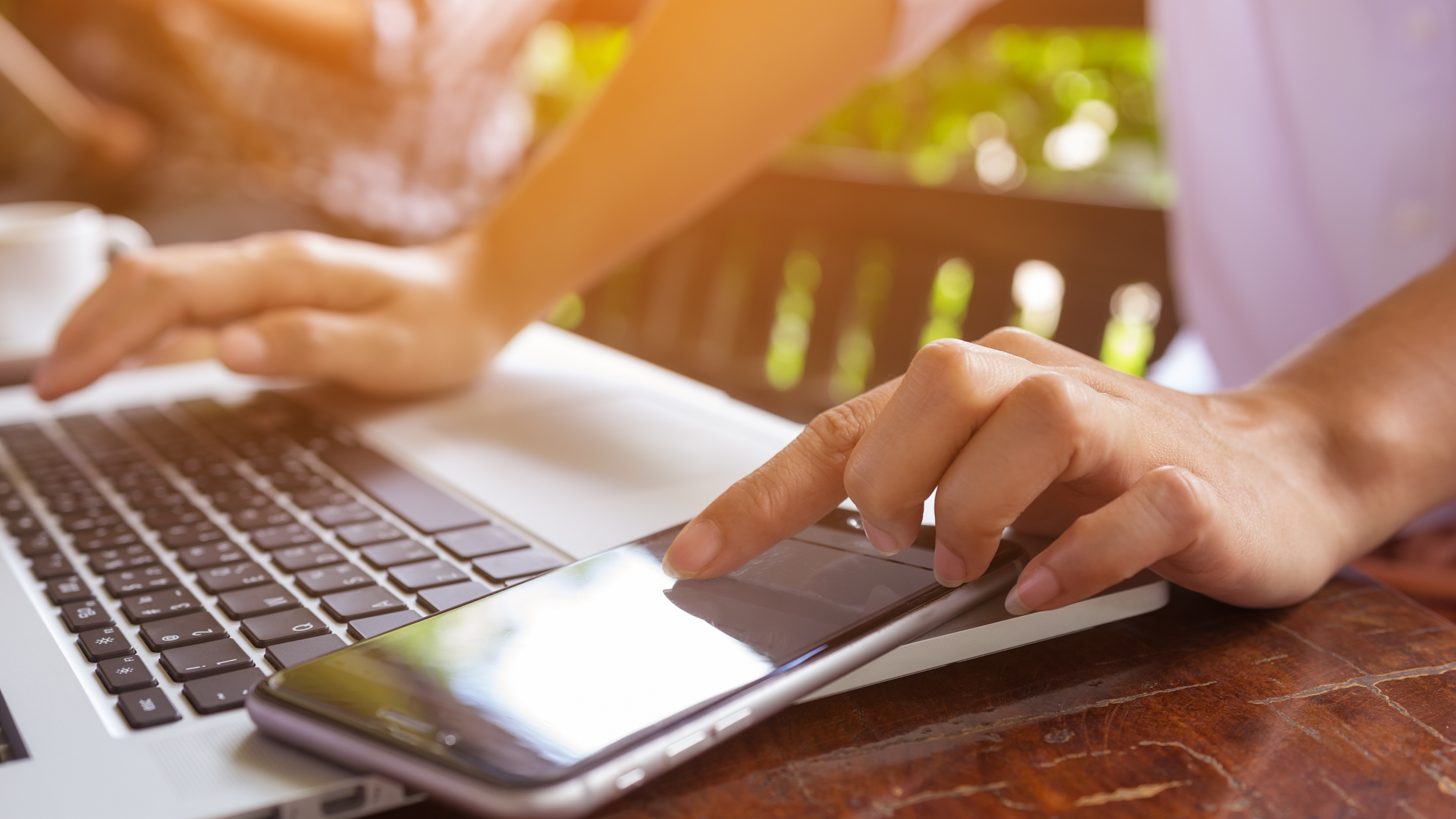 Close-up of a person using a smartphone and a laptop at a wooden table outside with blurred sunlight and greenery in the background.