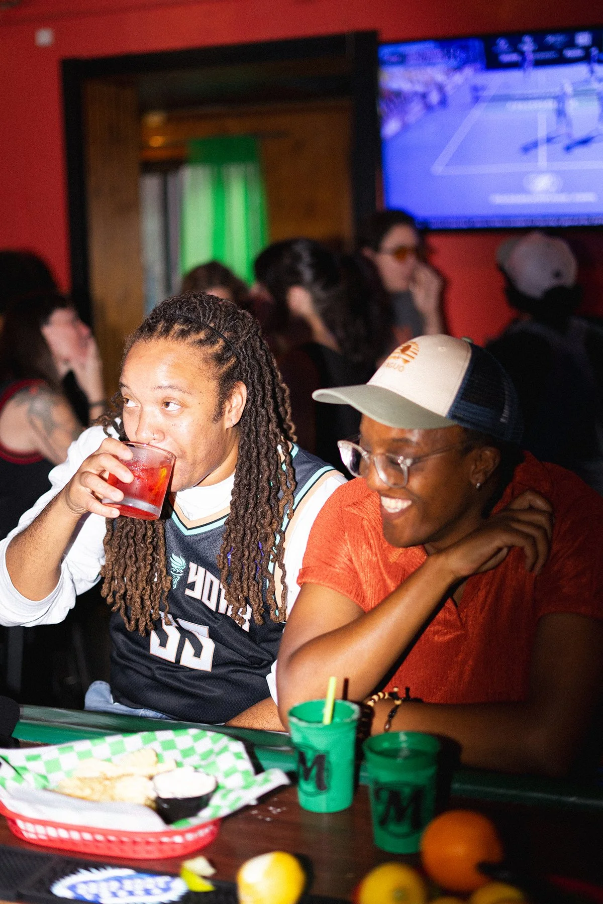 Two people smiling and drinking at Marsha's bar while watching a game on TV.