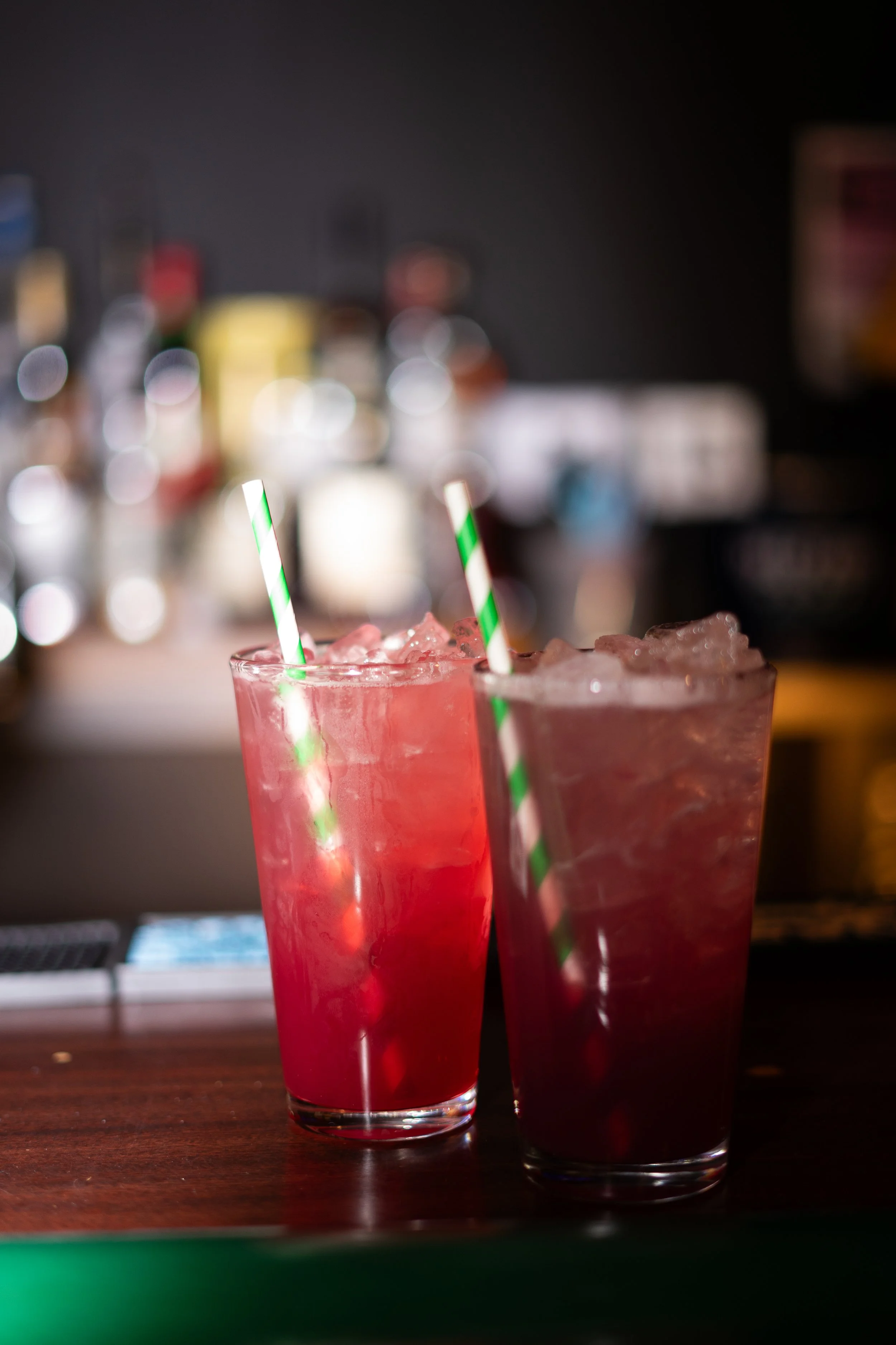 Two red cocktails with green and white straws on a wooden bar.