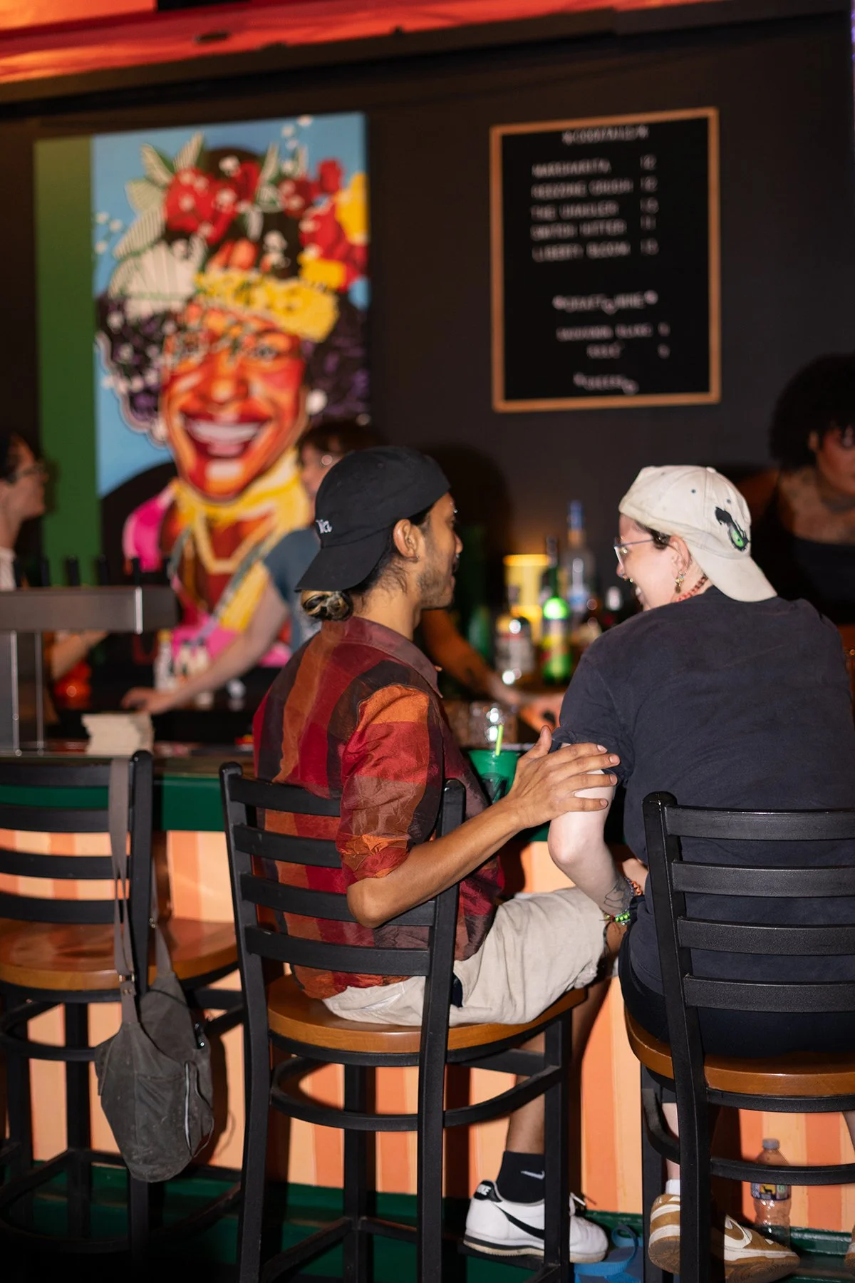 Two people sitting at the bar, in the background hangs a painted portrait of Marsha P. Johnson