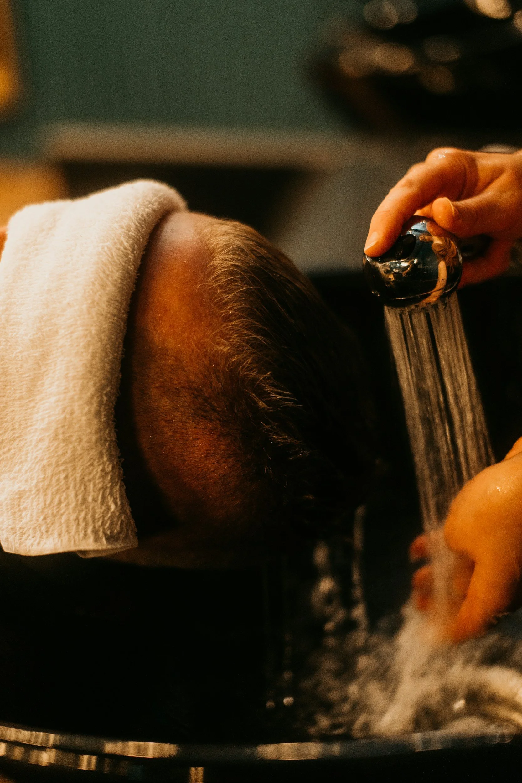 Person receiving a scalp massage under a handheld showerhead, with a towel draped over their head, in a spa setting.