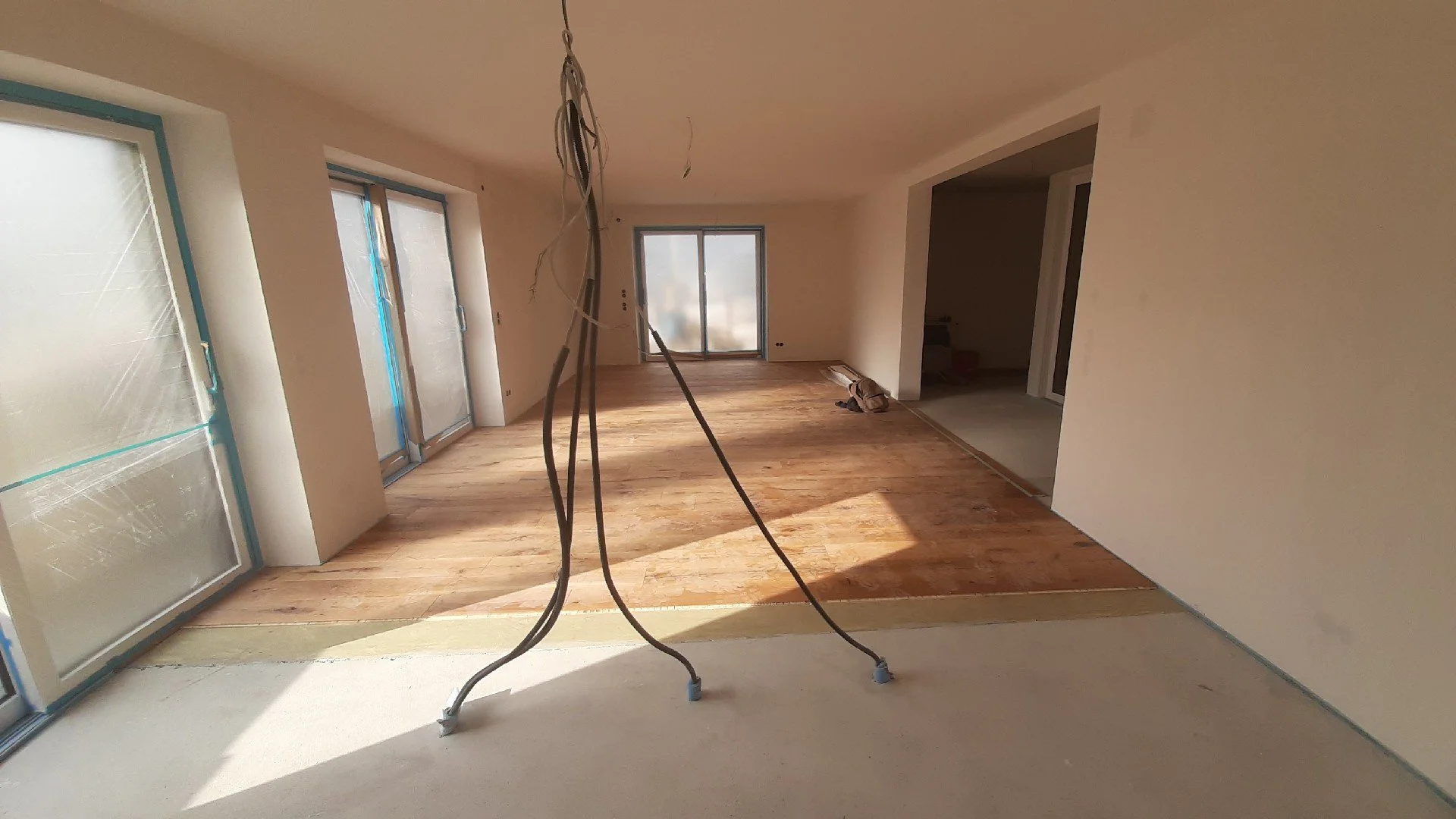 Innenansicht eines unfertigen Wohnzimmers mit weißen Wänden, Parkettboden und großen Fenstern, an denen noch Schutzfolie befestigt ist. Kabel hängen von der Decke.