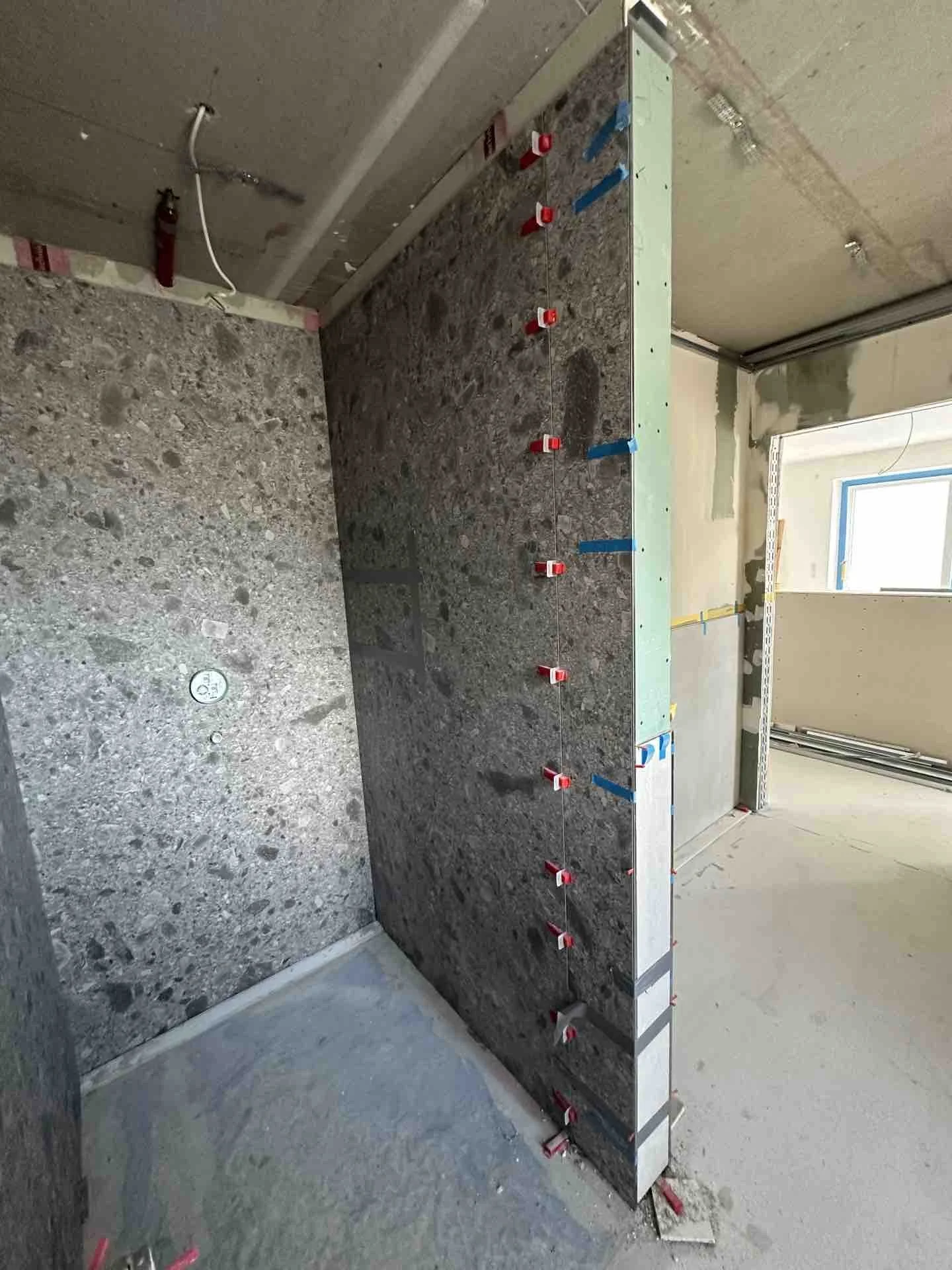 Bauarbeiten im Innenraum, eine Wand wird mit Fliesen oder Steinen verkleidet, mit Clippern für Fliesen oder Steine. Im Hintergrund eine ungeheure, unvollendete Wand mit Baumaterialien.