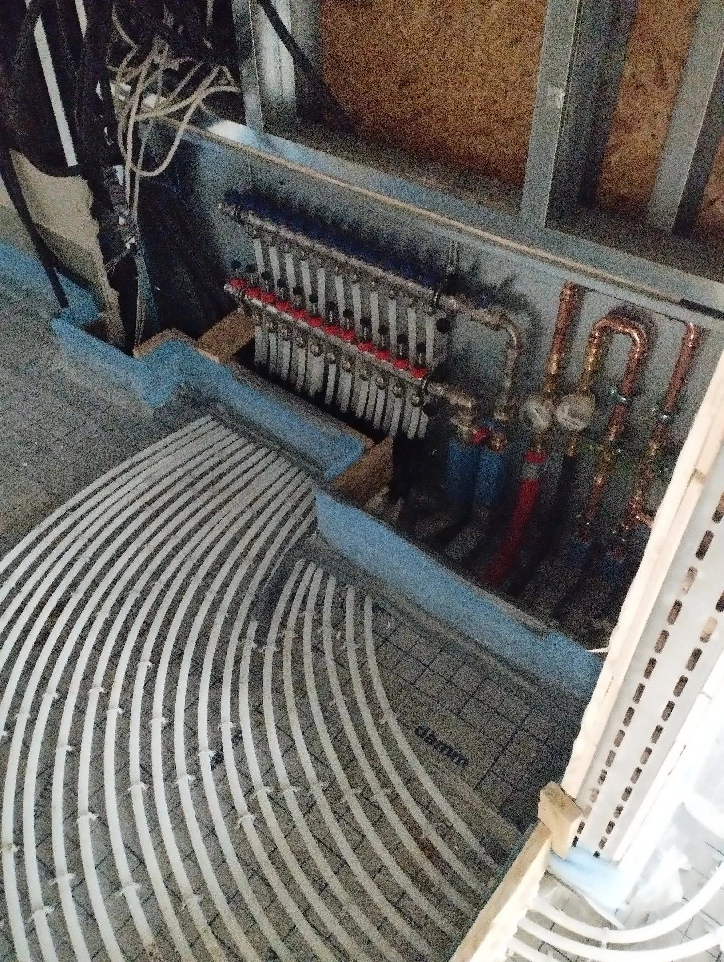 Installationsansicht eines Heizsystems mit Rohrleitungen, Ventilen, Messgeräten und einer Rohrmatte auf dem Boden, vermutlich im Bau oder in der Wartung.
