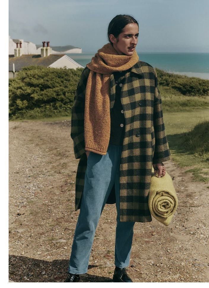 Woman standing outdoors on a dirt path with coastal background, wearing a long plaid coat, mustard scarf, blue pants, black shoes, and holding a rolled yellow blanket.