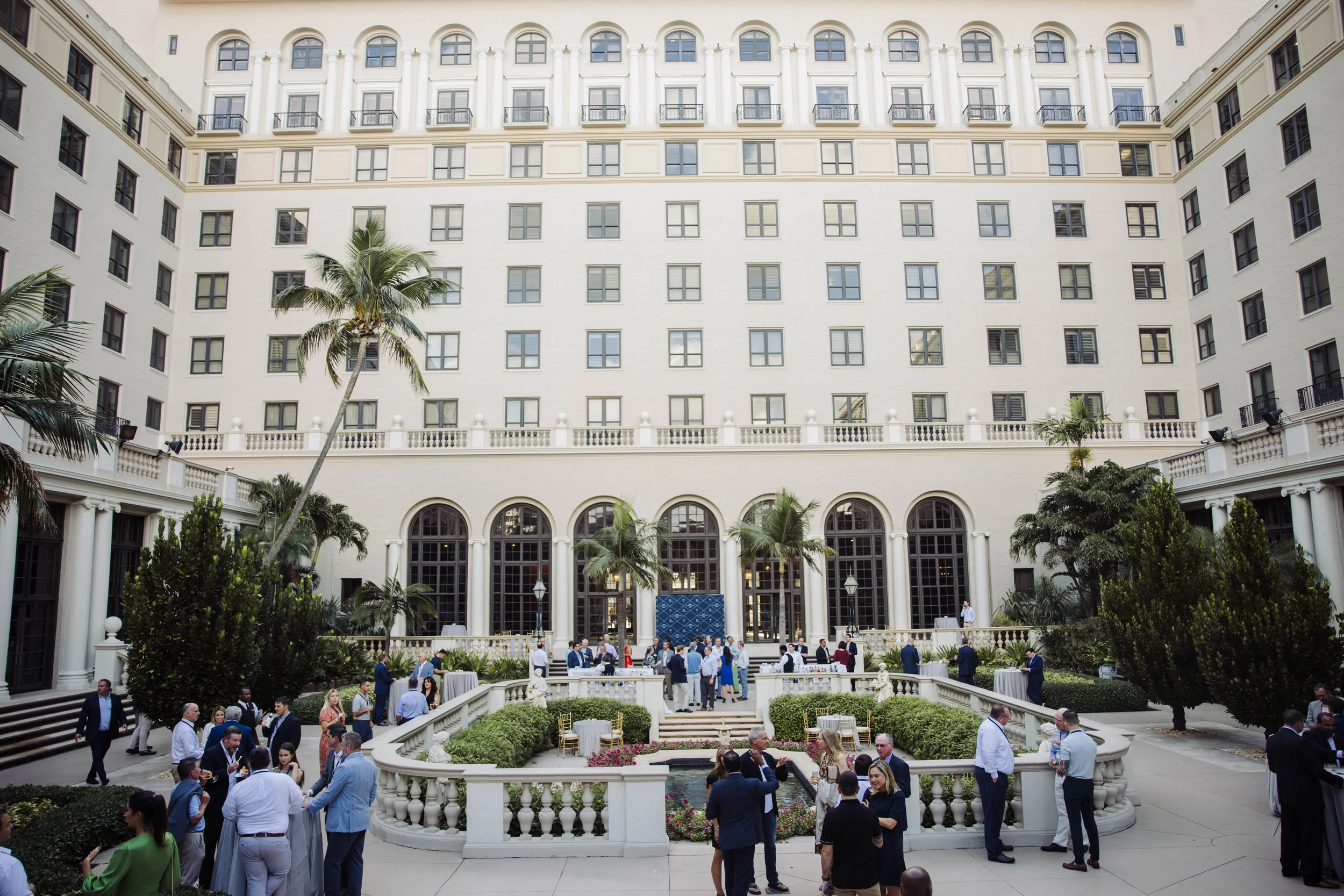 A hotel courtyard with people socializing, surrounded by tall palm trees, greenery, and white arches at The Breakers Palm Beach. The hotel building is in the background with multiple windows and balconies. Corporate event photography.