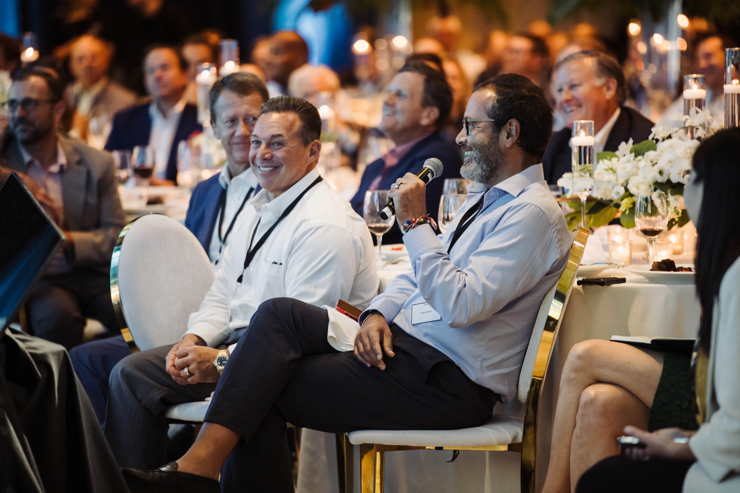 A group of people attending a conference or event, seated at tables with candles and floral centerpieces, with one man holding a microphone and smiling. The Breakers Palm Beach, corporate event photography