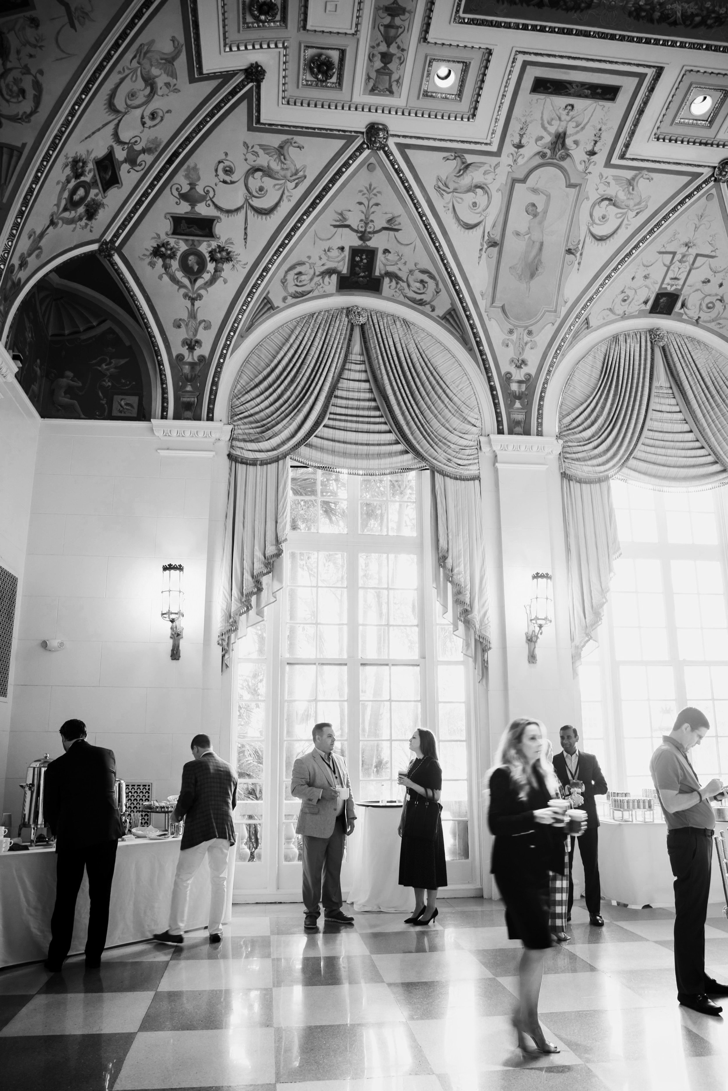 Interior of an elegant hall with ornate ceiling at The Breakers Palm Beach, large windows with draped curtains, and guests mingling at a social event.