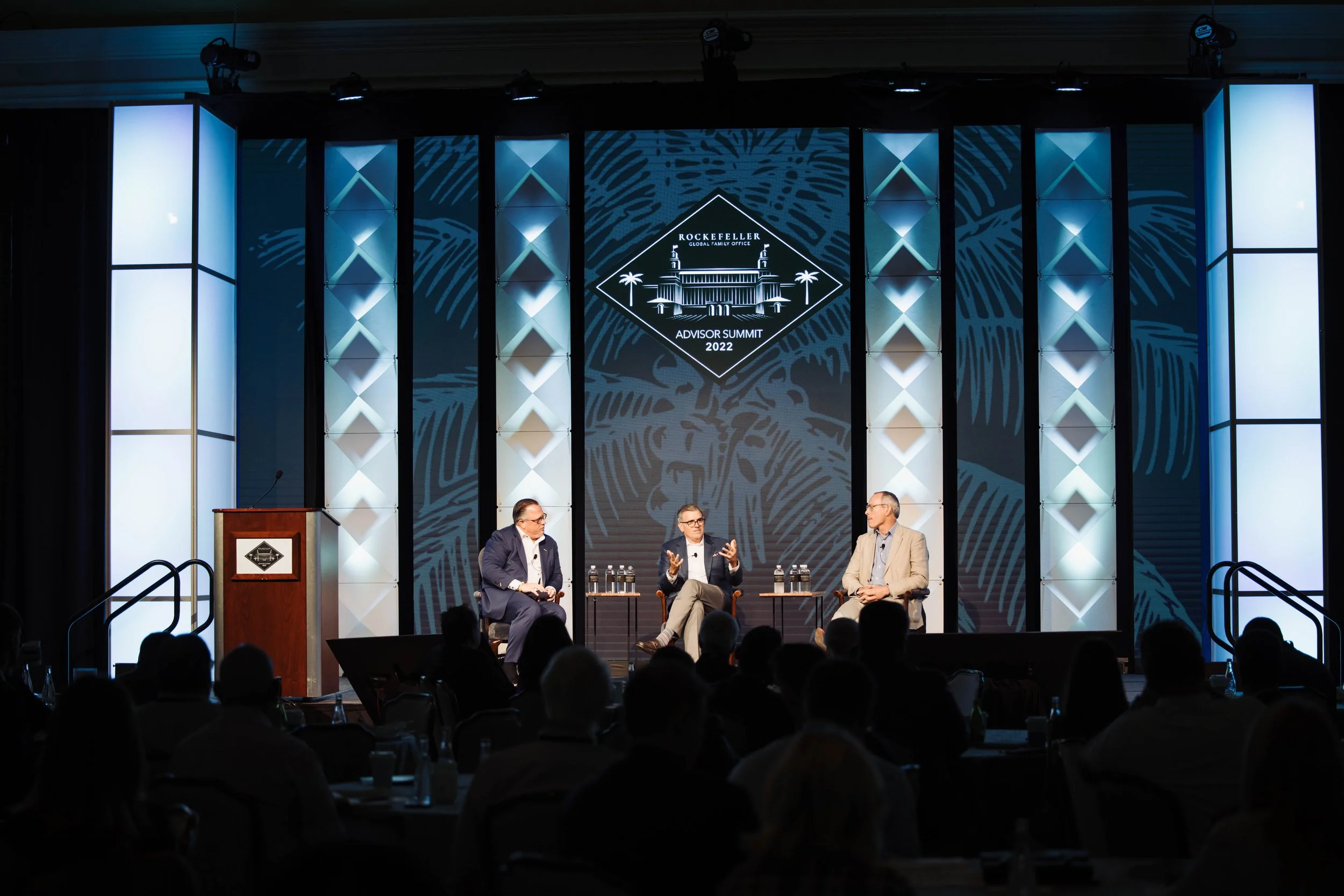 Three men seated on a stage during a panel discussion with a large screen in the background displaying the event's logo and a palm leaf pattern, in front of an audience in a conference hall at The Breakers Palm Beach.