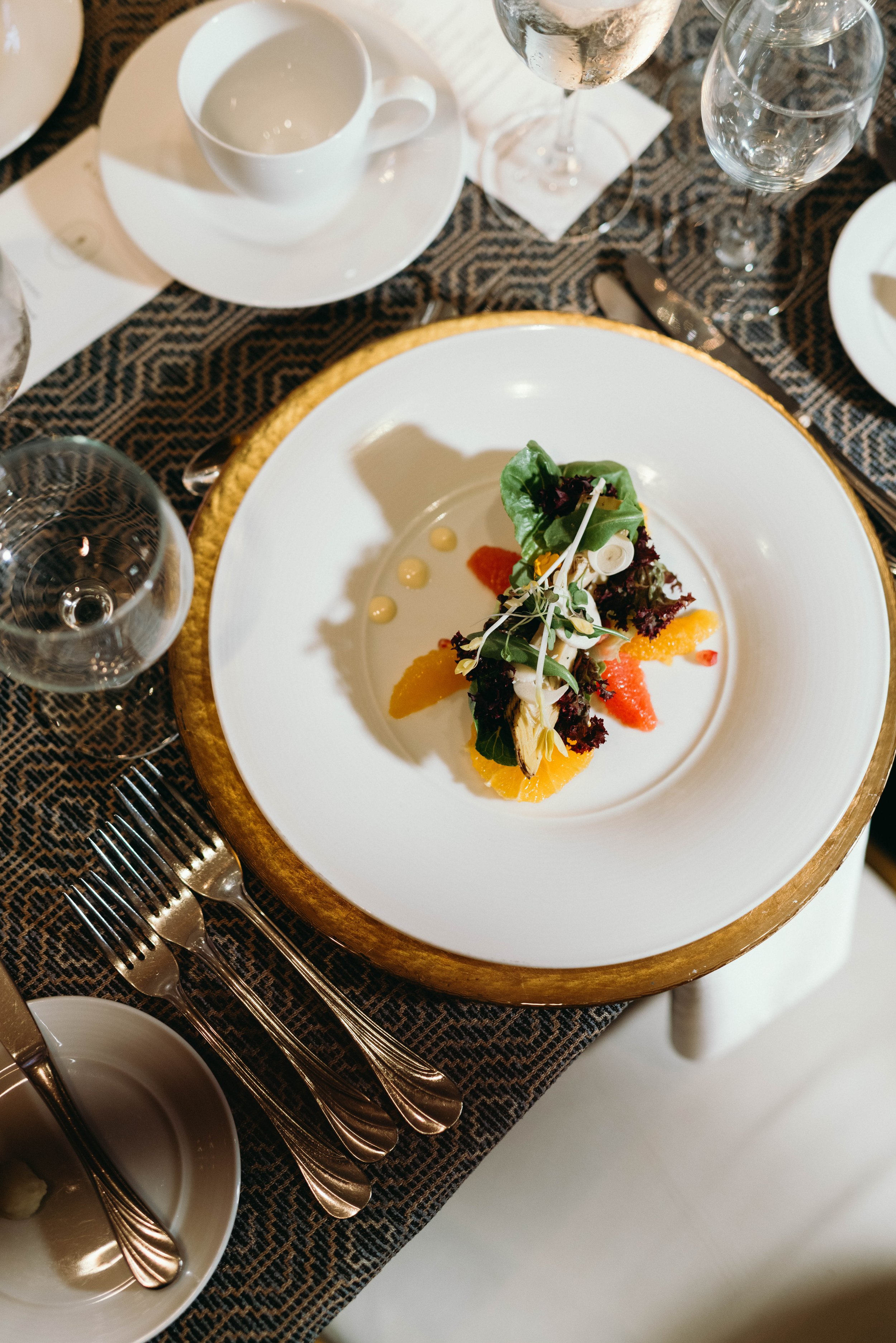 A gourmet salad with mixed greens, orange slices, and dressing droplets on a white plate with a gold rim, set on a black textured tablecloth surrounded by glassware, silverware, and a teacup.