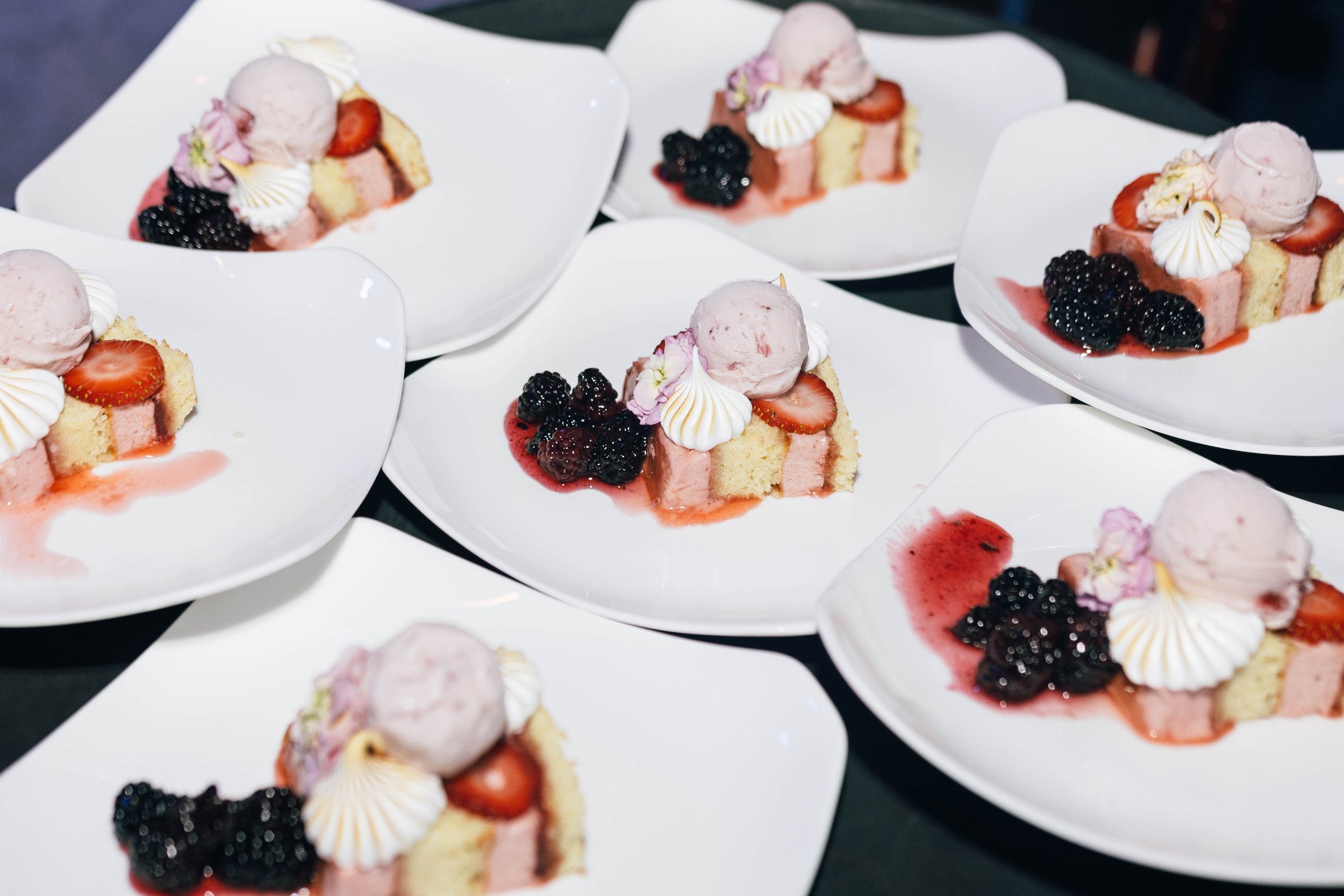 Multiple white plates, each with a slice of layered cake topped with pink ice cream, sliced strawberries, blackberries, a dollop of whipped cream, and small flowers.