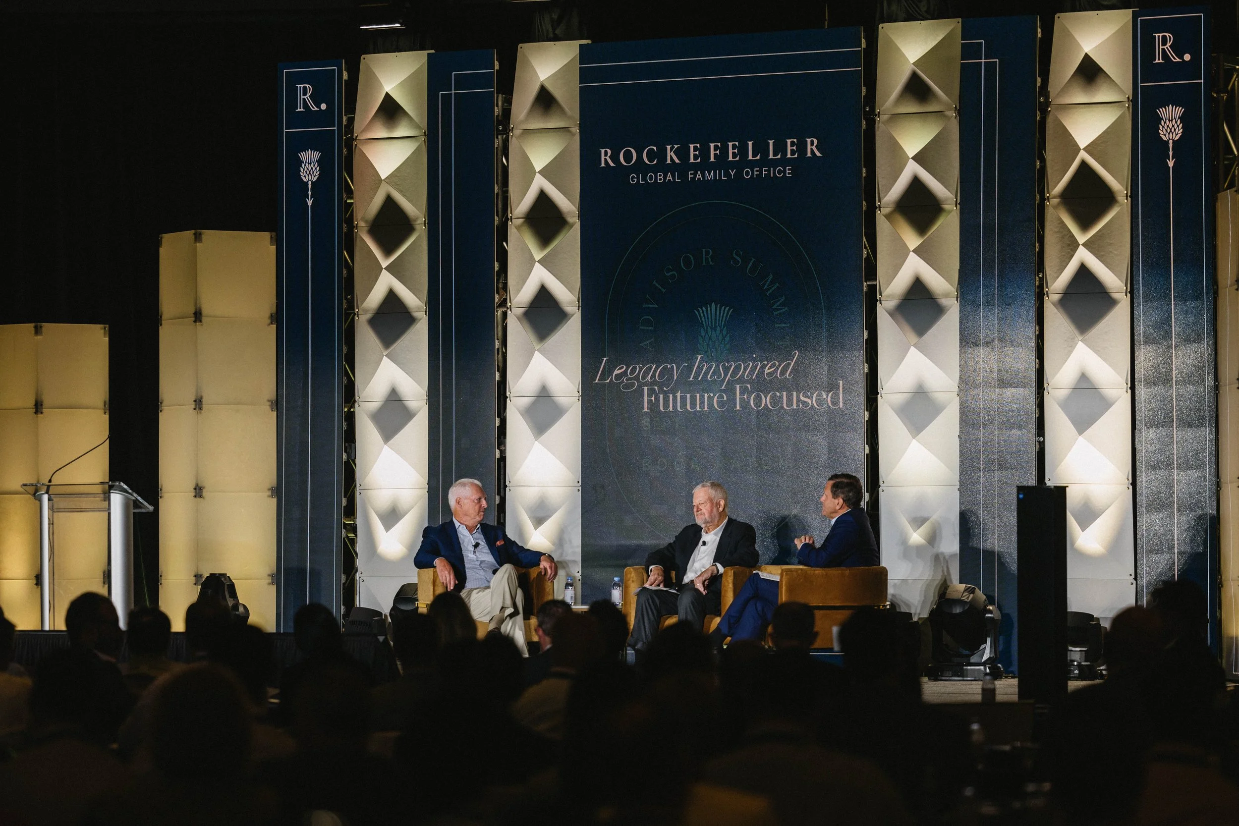 Three men are seated on stage in front of an audience at a Rockefeller event, with a large dark blue banner reading 'ROCKEFELLER GLOBAL FAMILY OFFICE' and 'Legacy Inspired Future Focused' behind them at The Boca Raton Hotel.