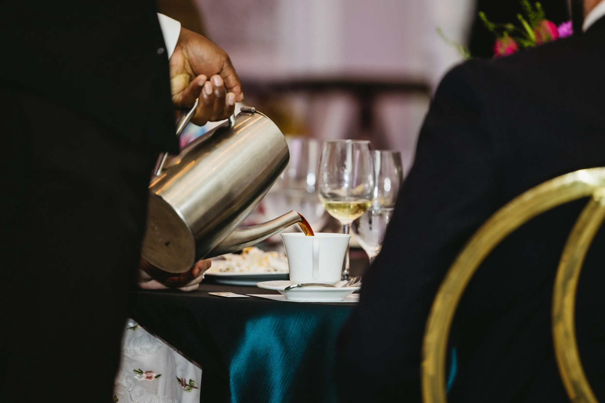 A person pouring coffee from a stainless steel pot into a white cup at a formal dining table with wine glasses and a woman with flowers at The Breakers Palm Beach.