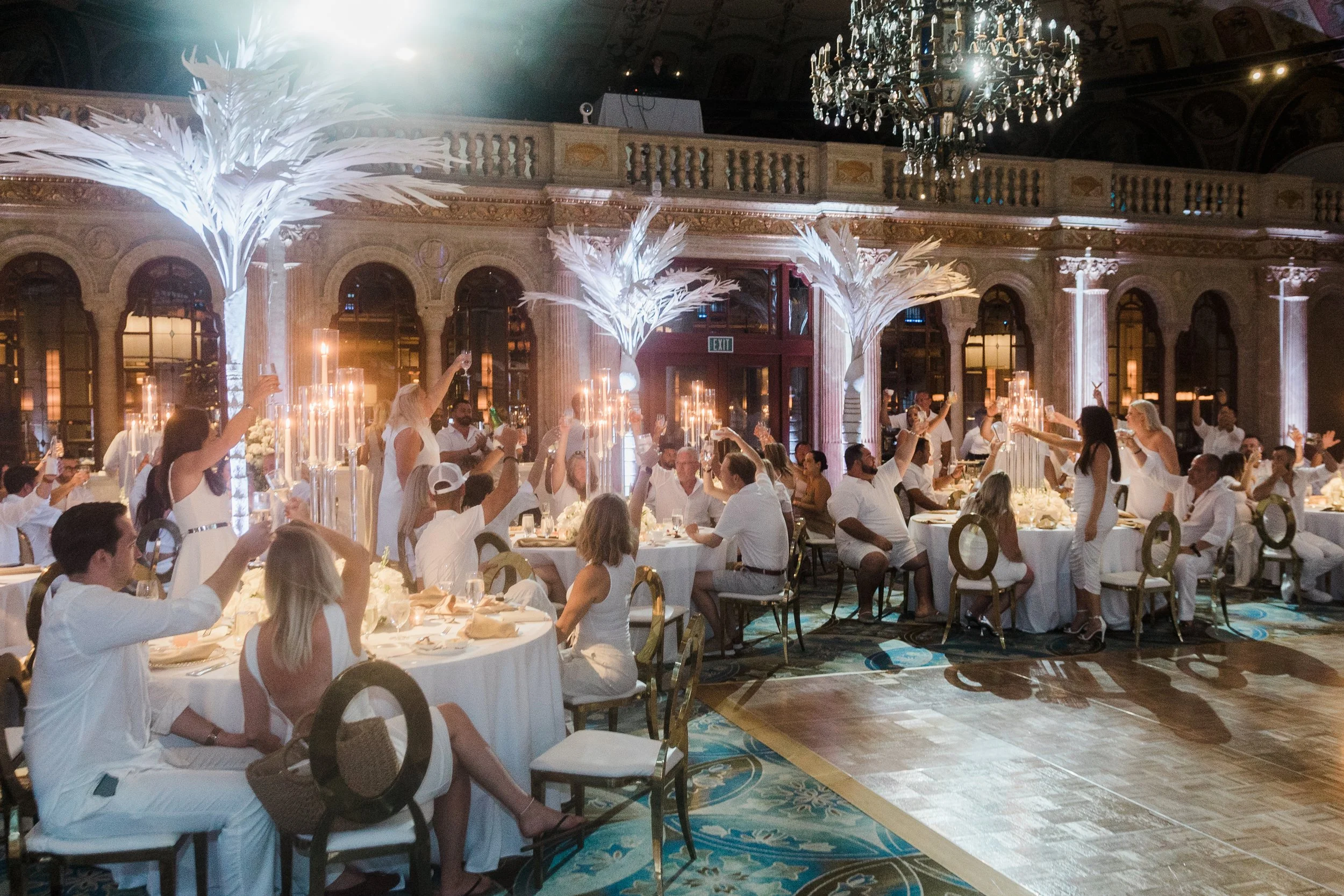 People celebrating during a corporate event, with tables decorated with candles and tall white feathered centerpieces, illuminated by a large chandelier, in an ornate room with high ceilings, large windows, and decorative columns at The Breakers.