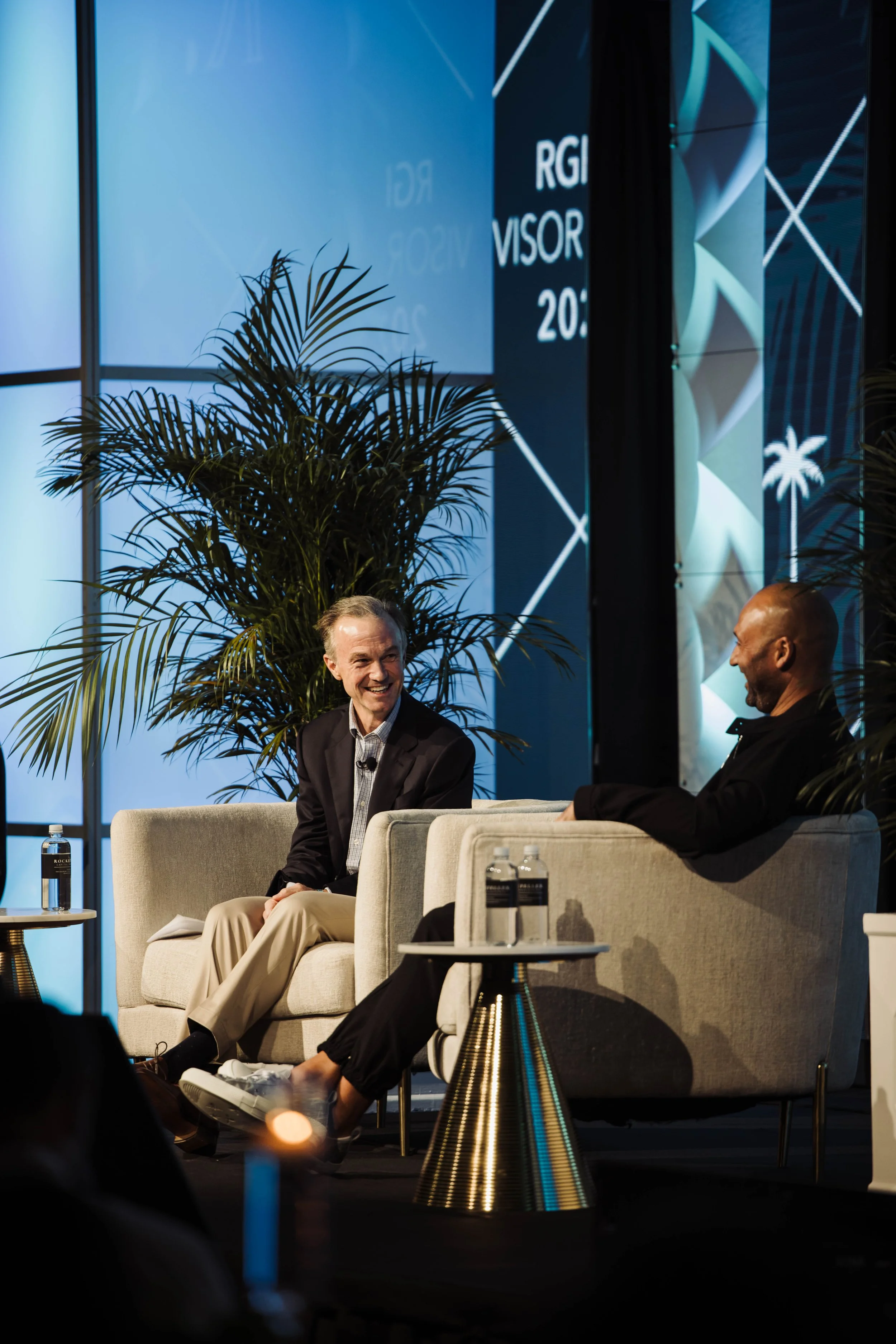 Two men having a conversation on stage at a conference, sitting in beige armchairs with small side tables holding water bottles, with posters and a large plant in the background. The Breakers Palm Beach