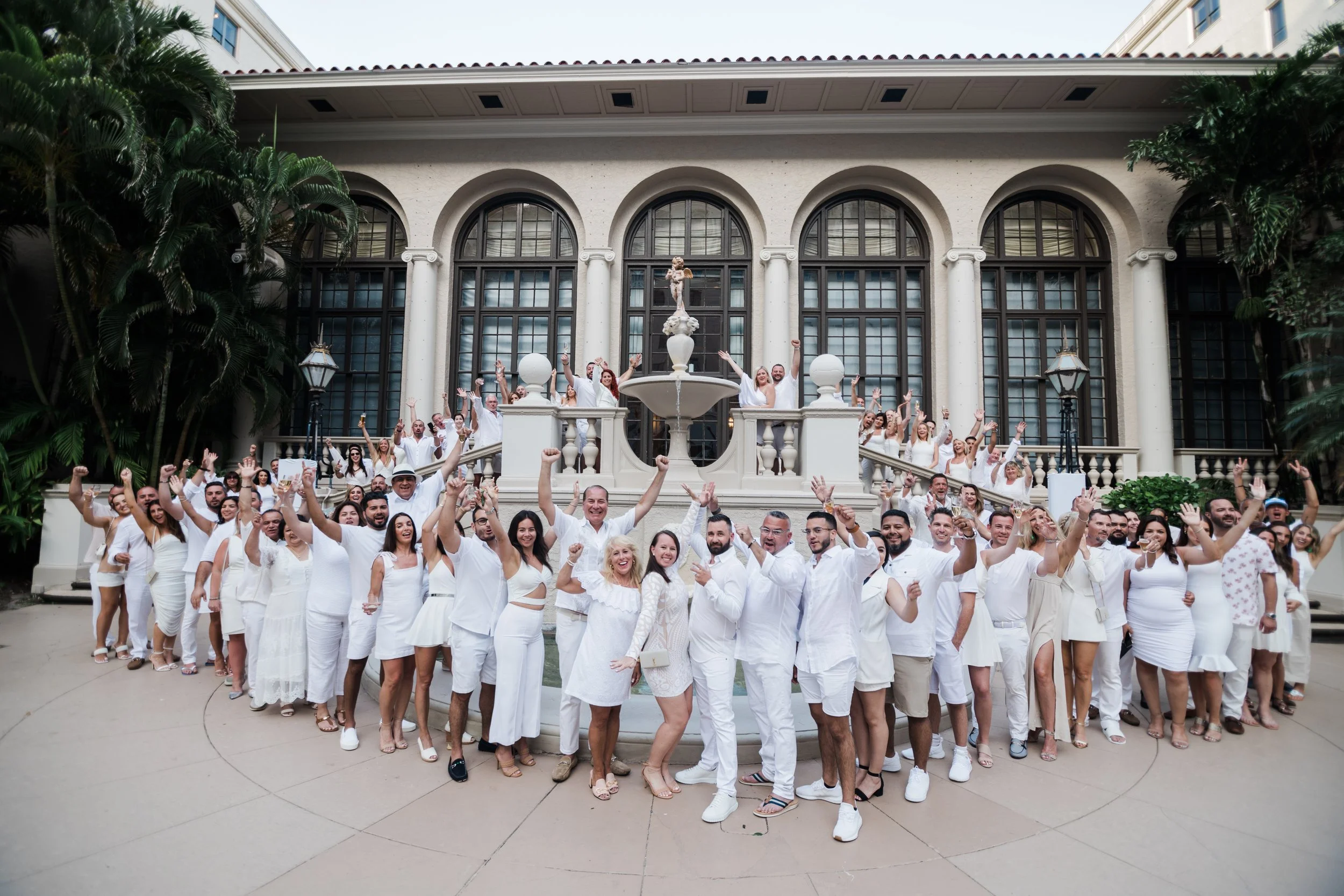 Group of people dressed in white celebrating in front of a historic building at The Breakers Palm Beach with arched windows and a fountain. Corporate event photography.