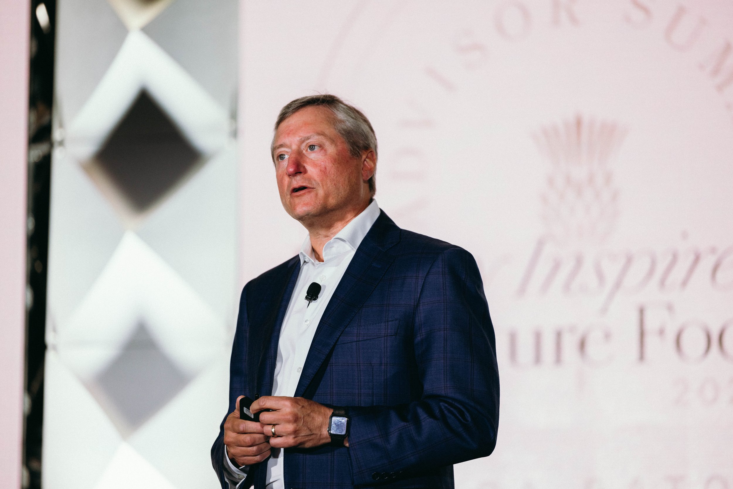A man in a dark suit holding a device, standing in front of a large screen with a blurry presentation or logo in the background. The Boca Raton Hotel, corporate event photography