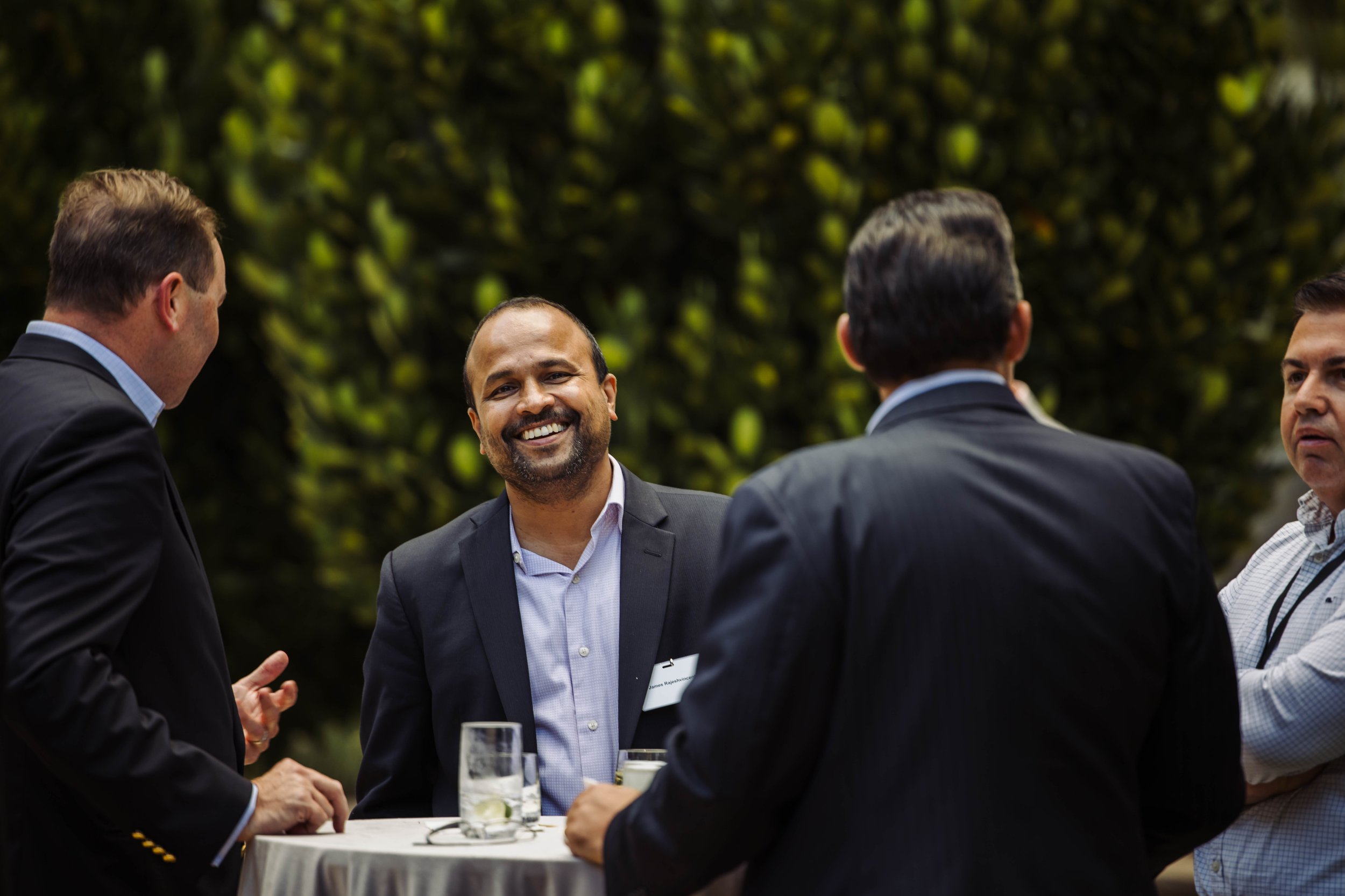 Group of four men in suits having a conversation outdoors around a small round table with drinks, smiling and engaging in a discussion. The Breakers Palm Beach, corporate event photography